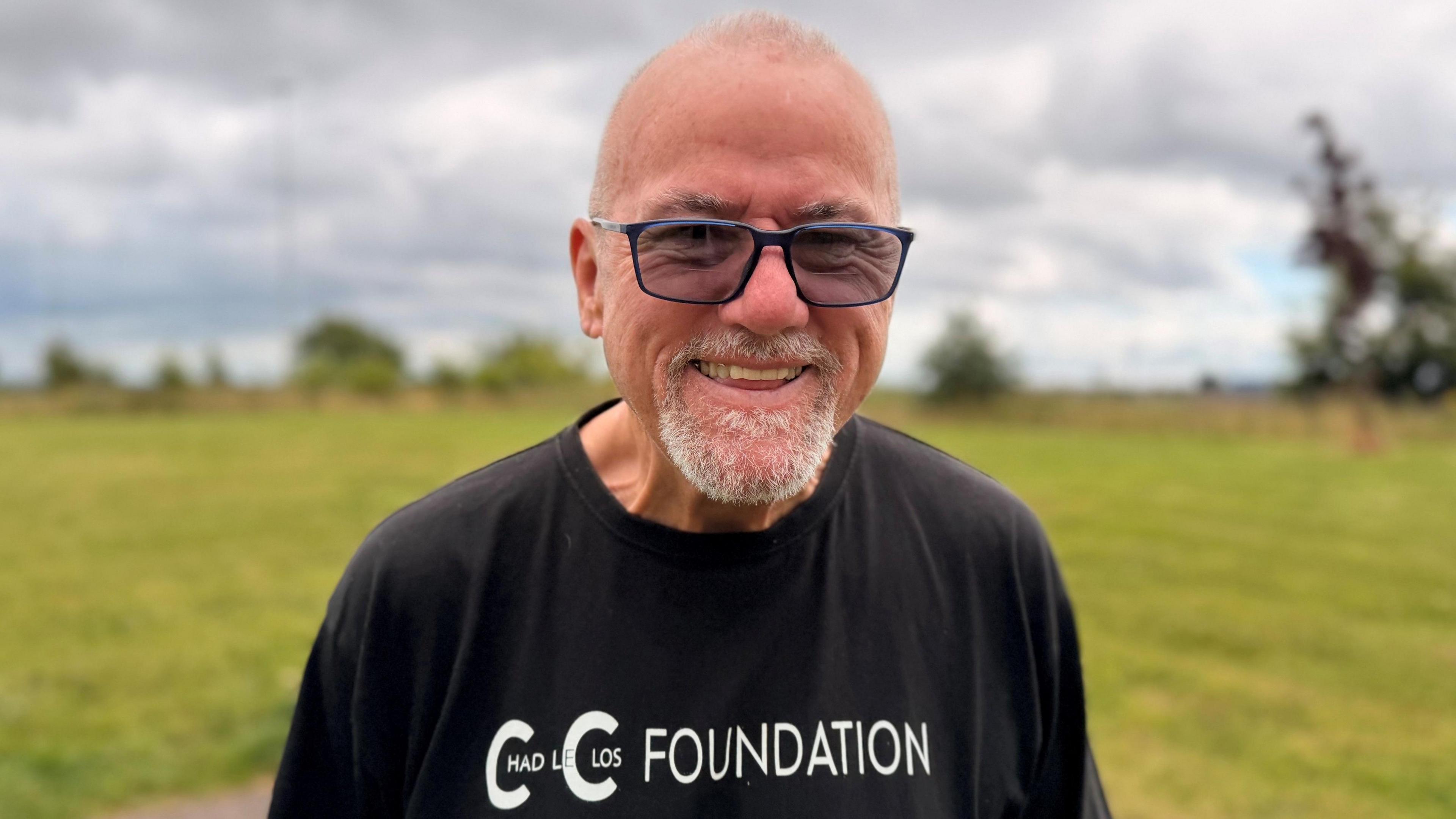 A smiling bald man with a grey goatee and dark-rimmed glasses. He is wearing a black Chad le Clos Foundation t-shirt