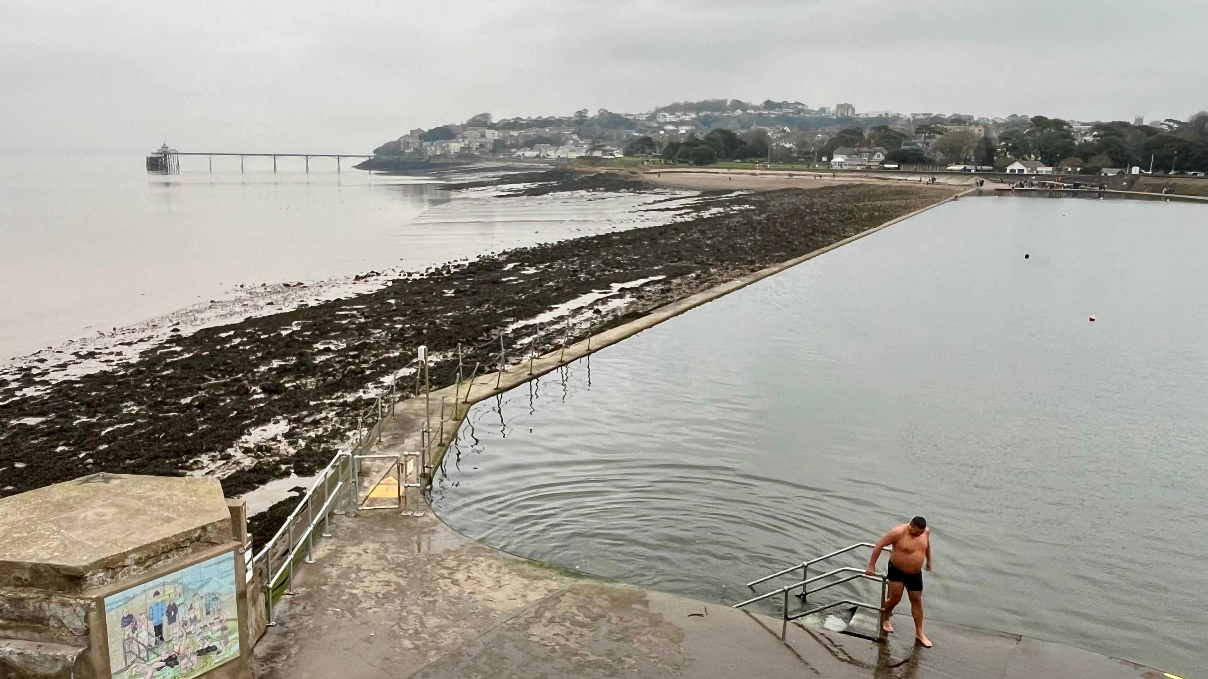 A man wearing swimming shorts gets out of Clevedon Marine Lake. In the distance the town and its pier and beach are visible. There is nobody else in the lake and there are ripples going out from the steps where the man has just climbed out 