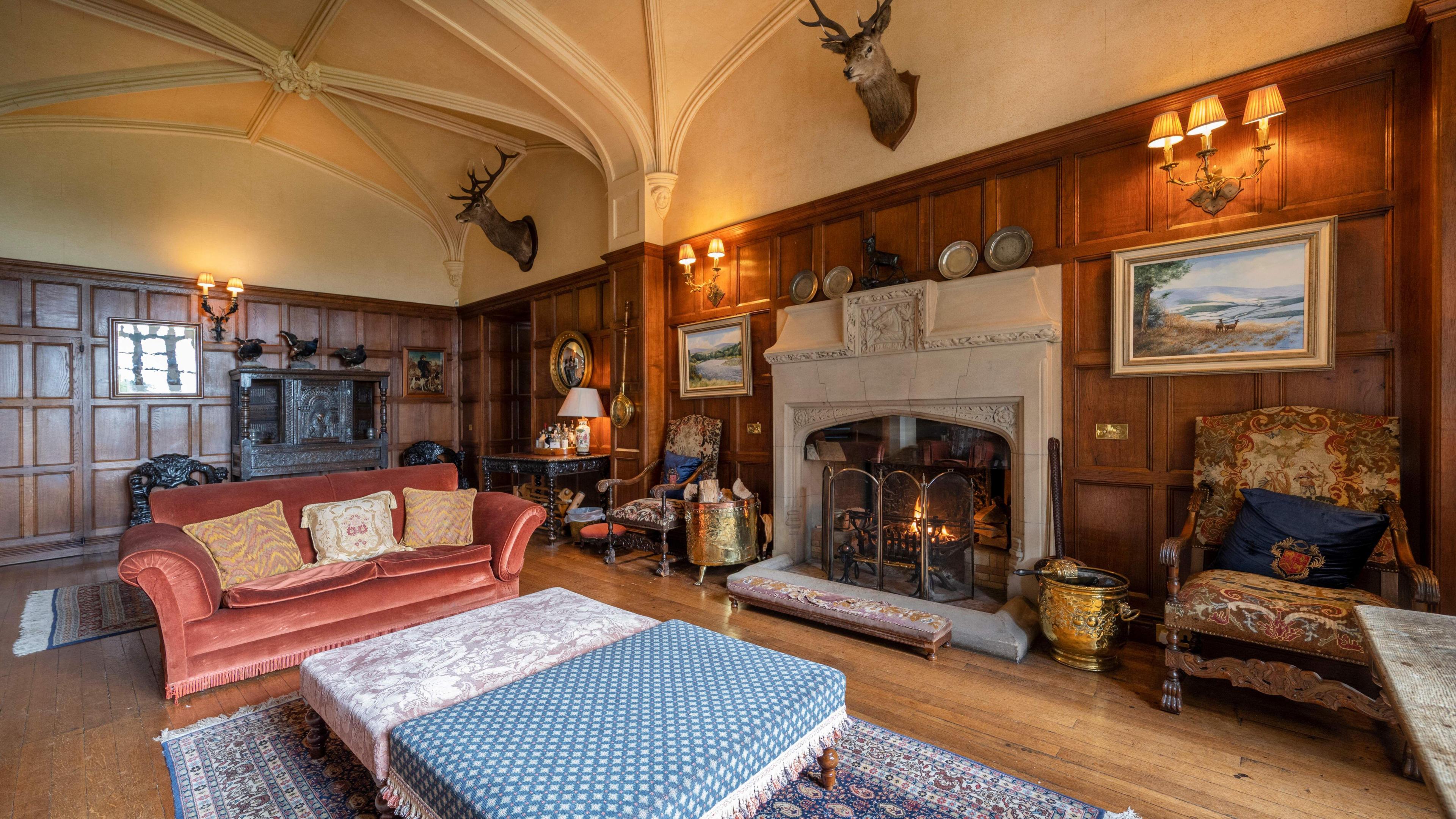 A 19th century stately living room with two stag heads on the walls, one above a large stone fire place. 