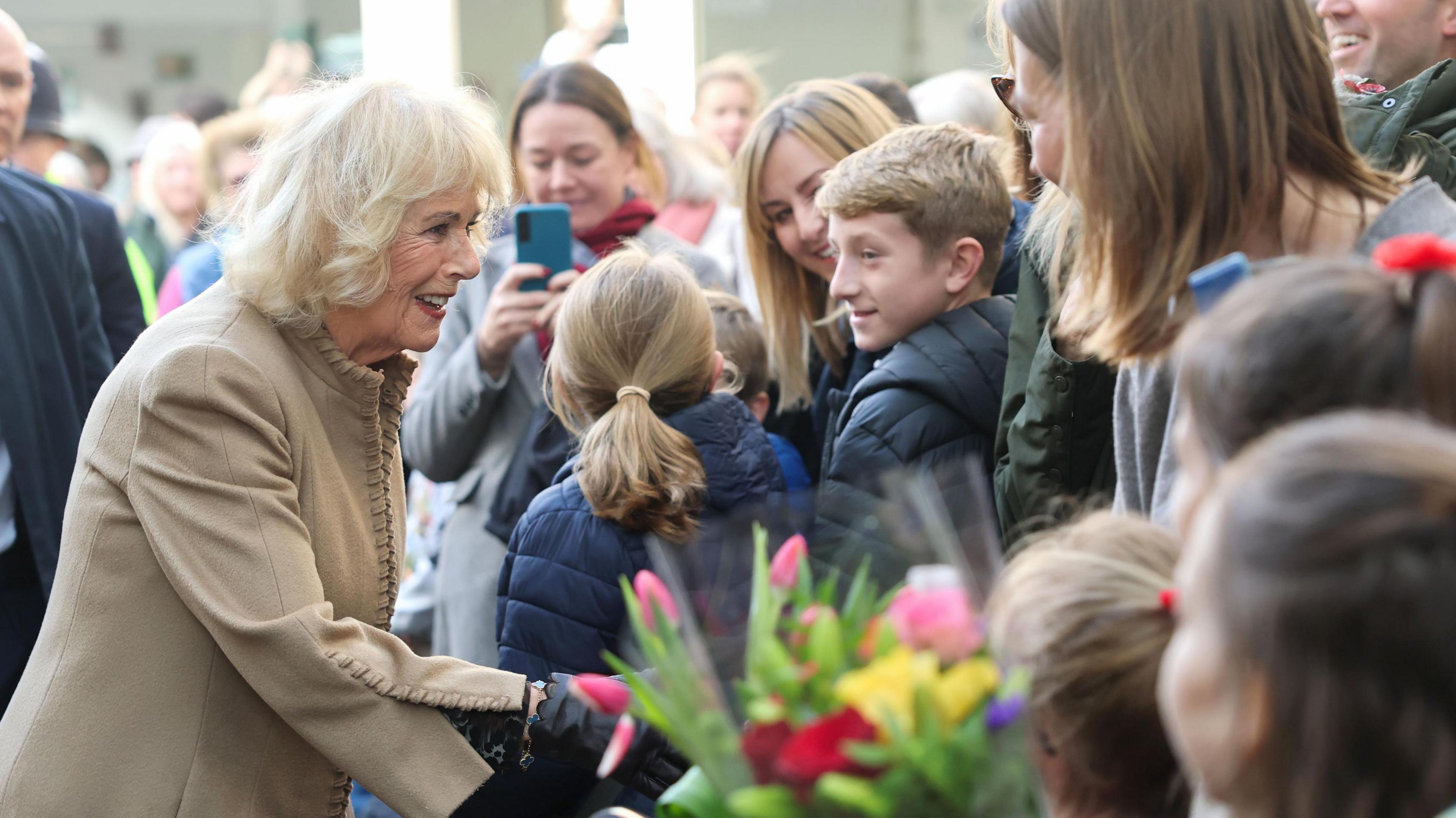 Queen Camilla wears a brown jacket and shakes hands with a well-wisher with people watching her behind a fence