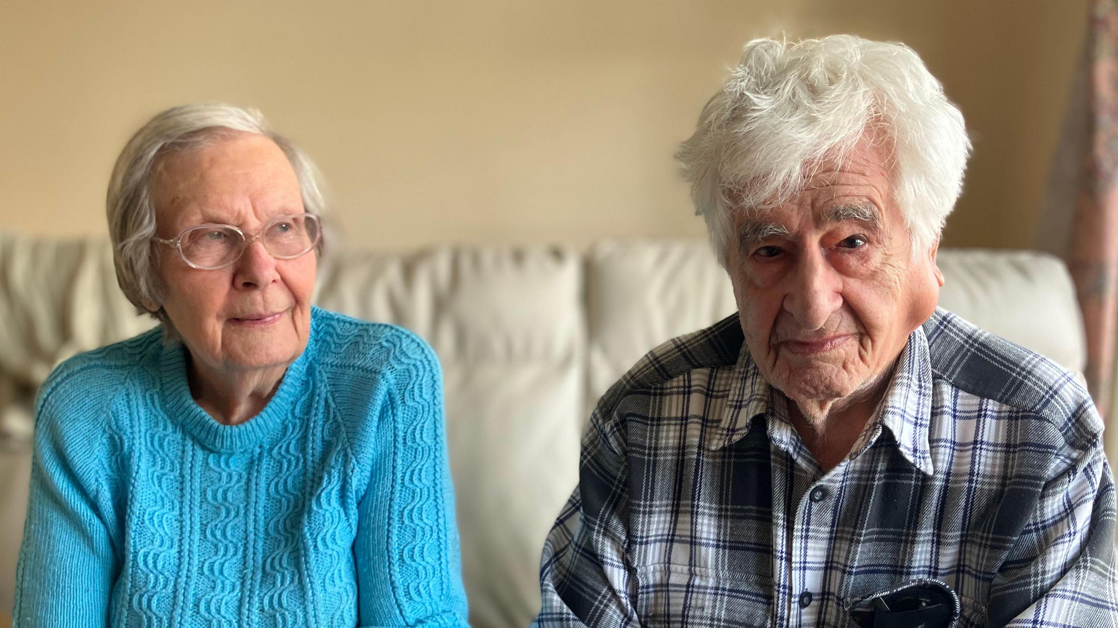 An elderly couple sat on a white sofa. The lady is wearing a blue jumper and the man is wearing a check shirt. Both look serious. 