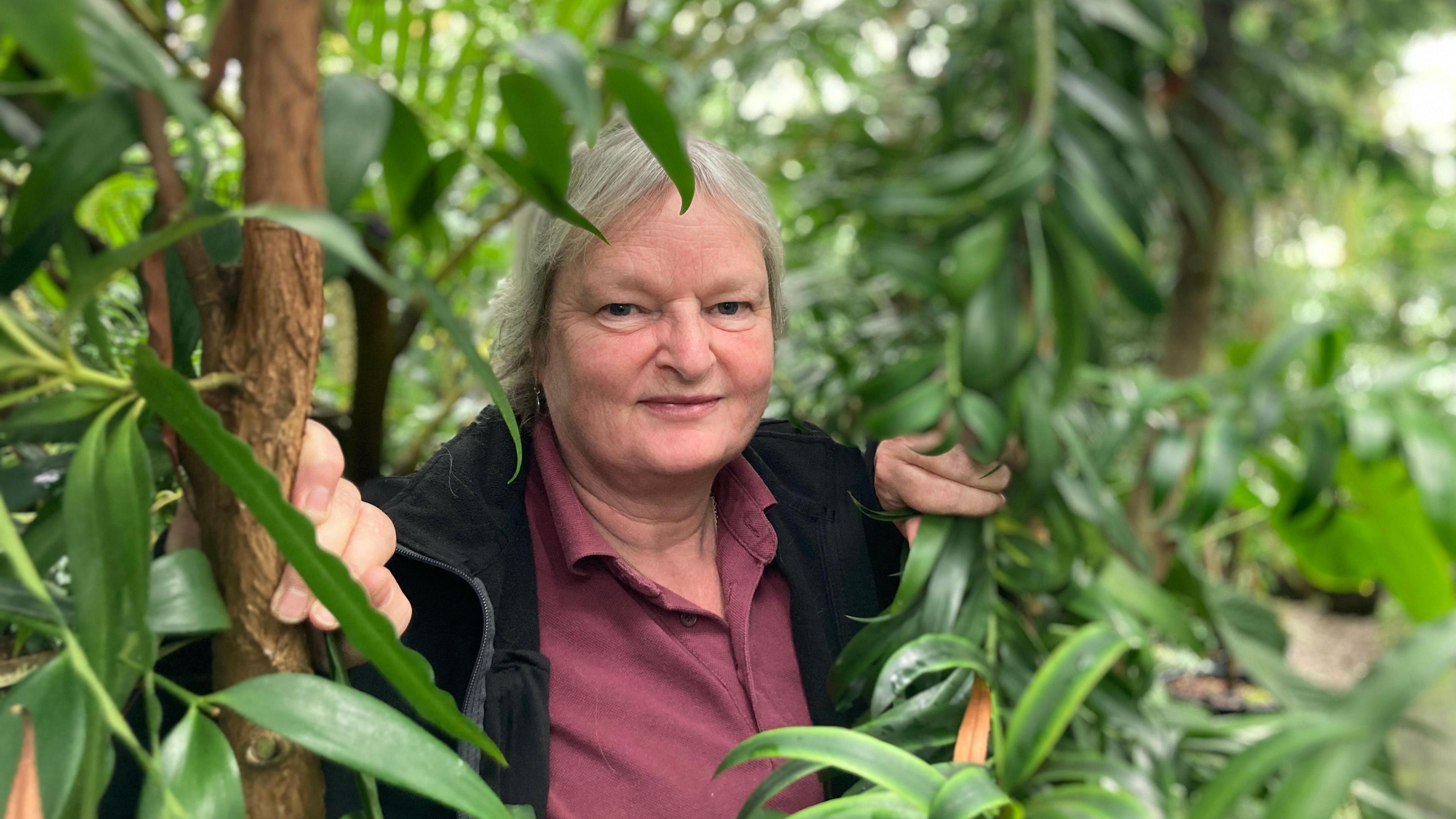 Fiona is pushing through some leaves in the botanic gardens. She has grey hair and a maroon top under a black jacket or cardigan.