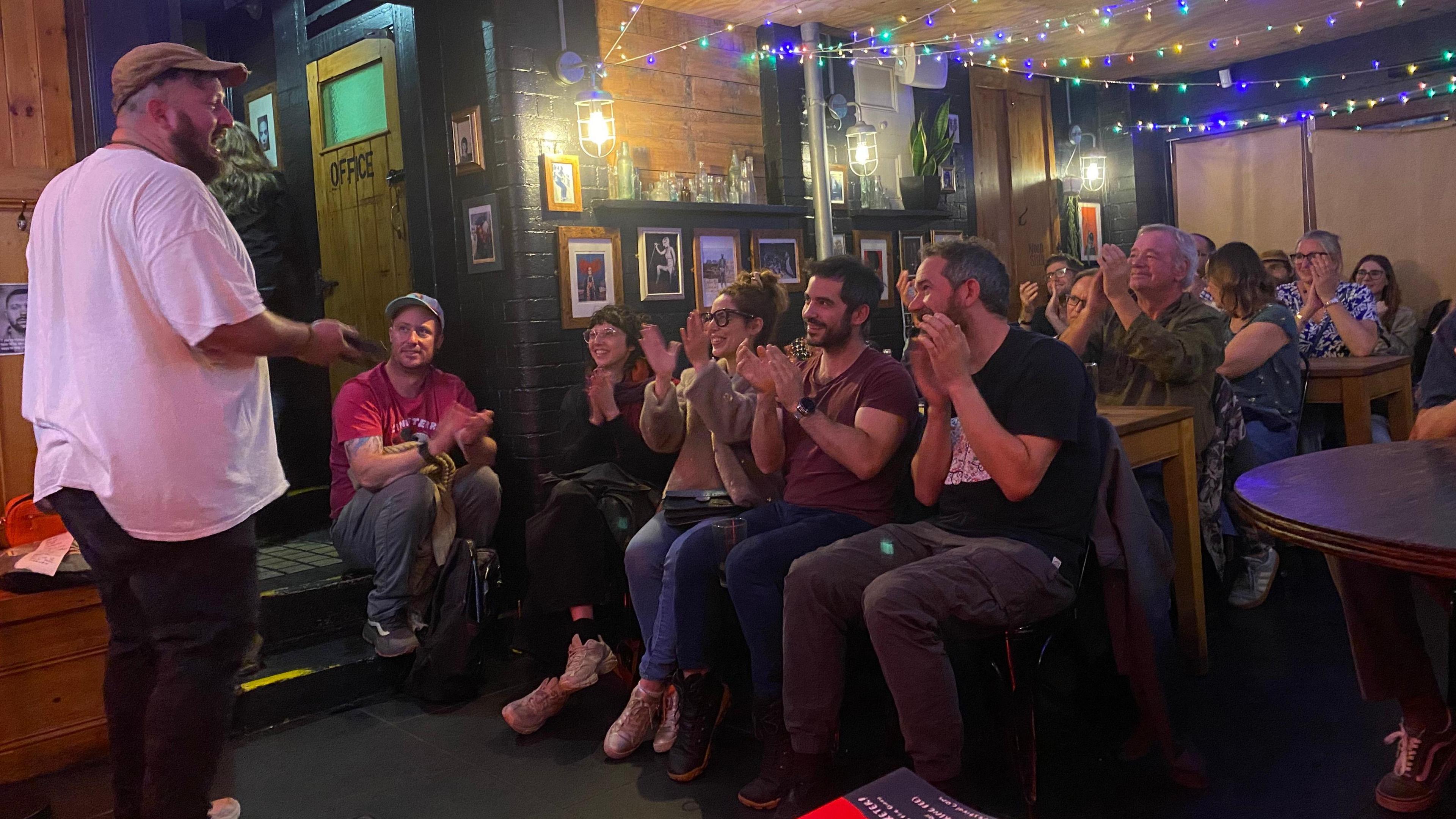 Kev Mud, a comedian in a white t-shirt, performs to a room of people who are applauding him