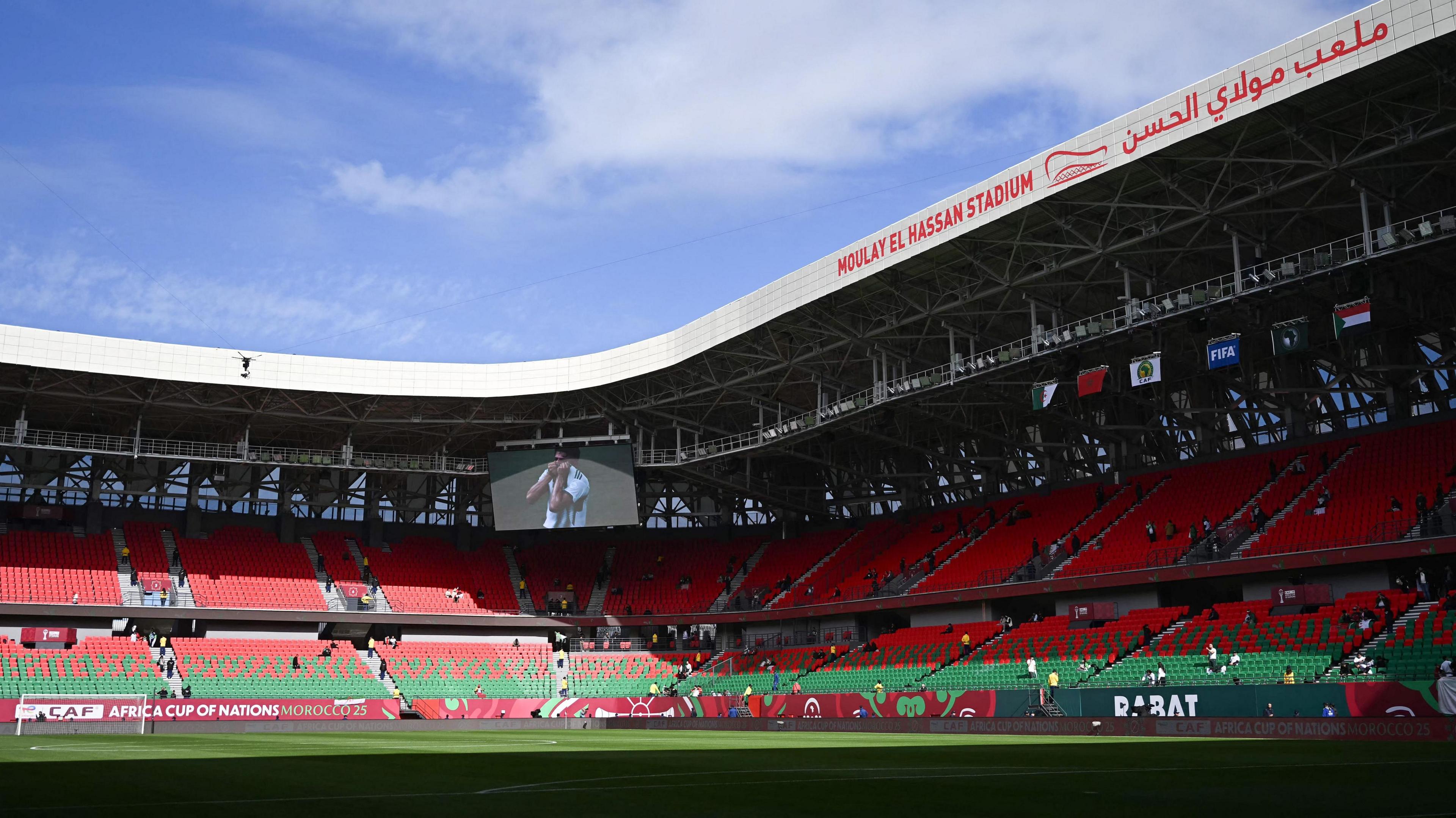 A general view of the inside of the Moulay Hassan Stadium in Rabat, looking towards one corner, showing red and green seats. The green pitch is partly in shadow as a blue sky with light cloud shines above the grey roof which arcs around and contains the name of the stadium in red English and Arabic text