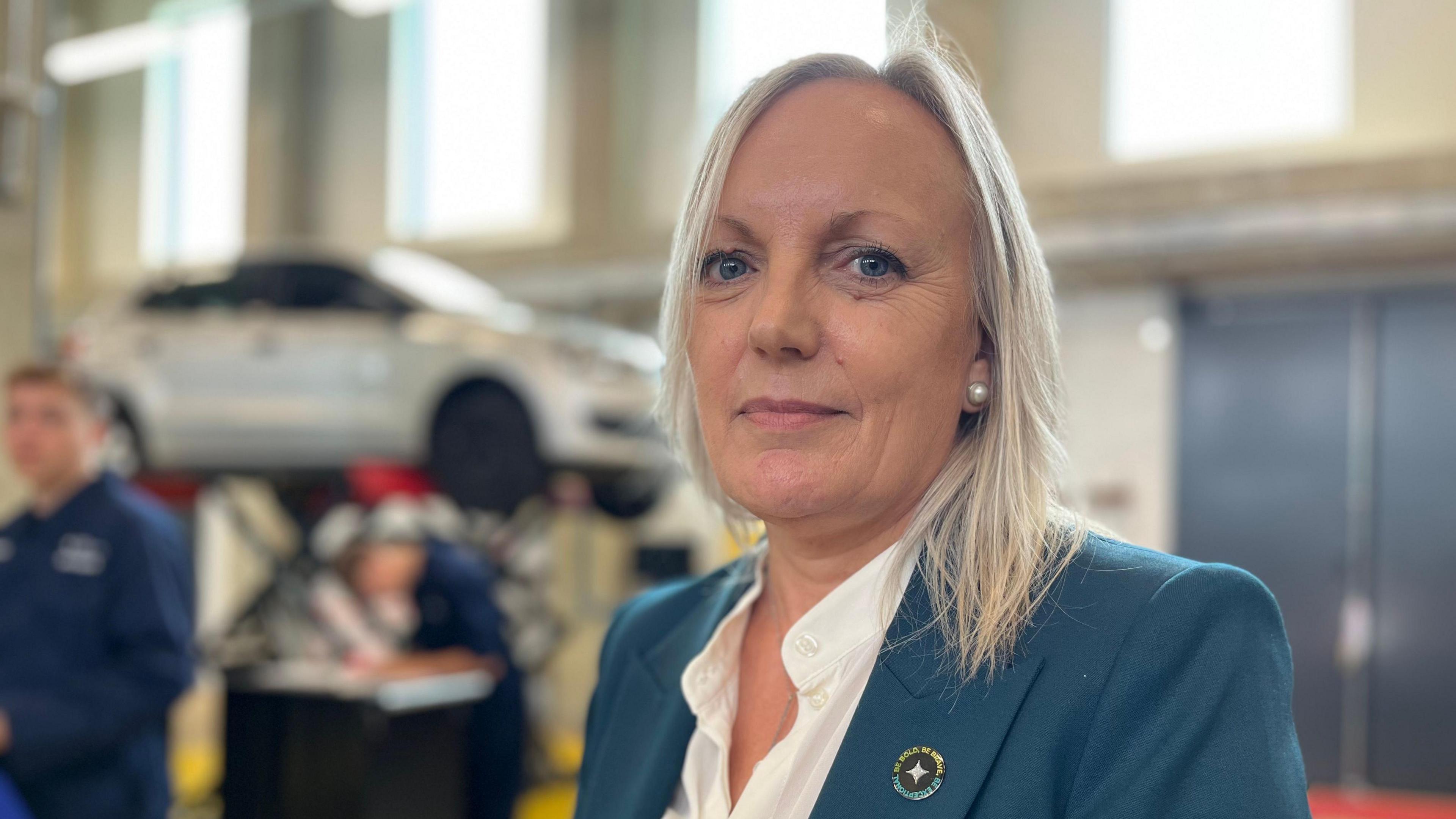 Julia Bates, who has with long blonde hair in a dark greeny/bue jacket. She is standing in a large building, with motor mechanic students behind her. 
