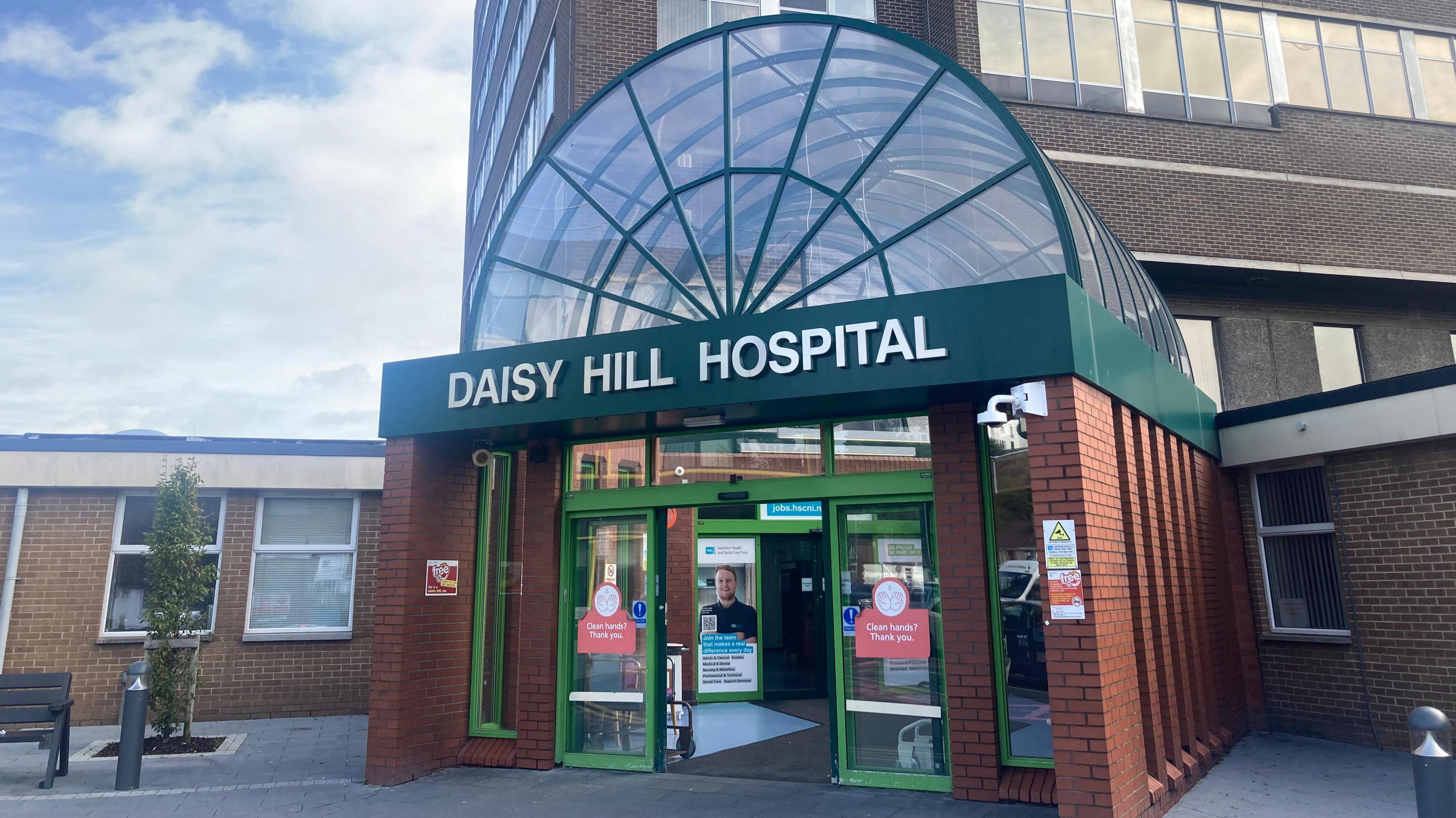 A large brown bricked building has a green banner on it's front that reads 'DAISY HILL HOSPITAL.' The banner sits at the entrance to the building below a glass archway which is encased in a green skeletal structure. They sit on-top of a red brick extension out the front of the brown bricked building. There is a black bench and a small green tree to the left of the entrance