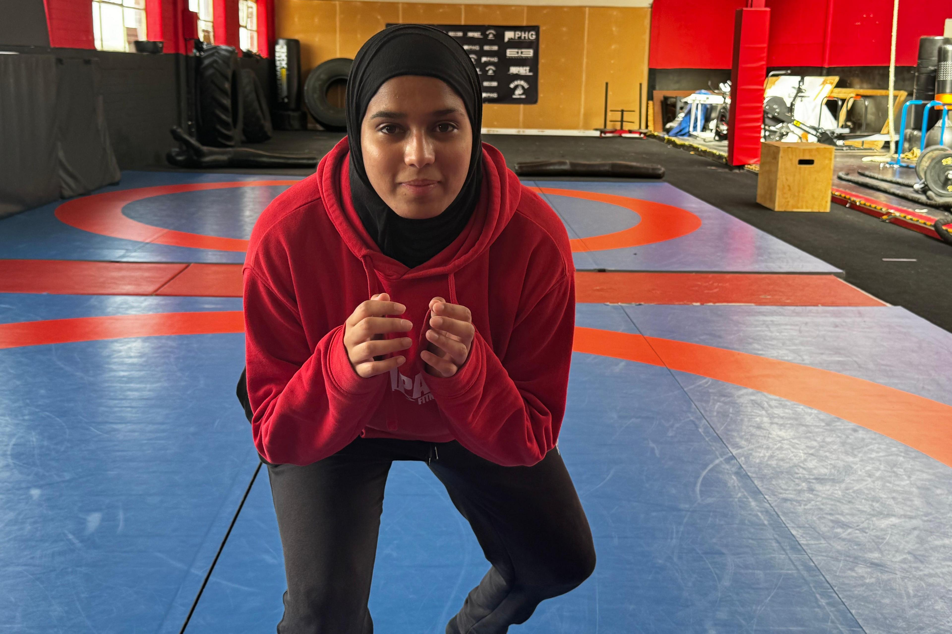 A woman wearing a black hijab, red hoodie, and black trousers squats in a wrestling position, in a room at the wrestling club with a blue and red floor