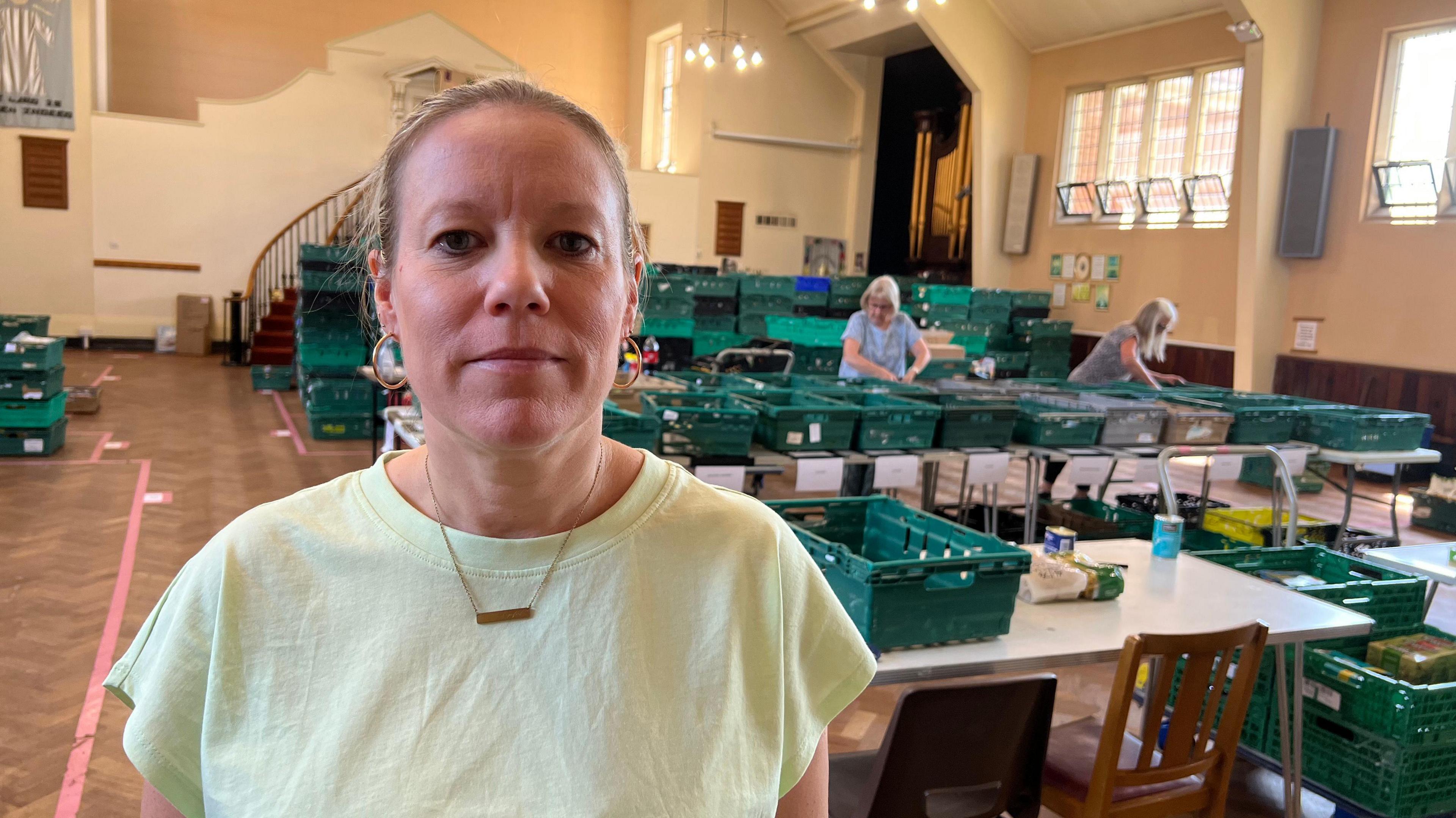 A woman is looking at the camera standing in front of rows of tables inside a large hall, with green boxes on all of them.