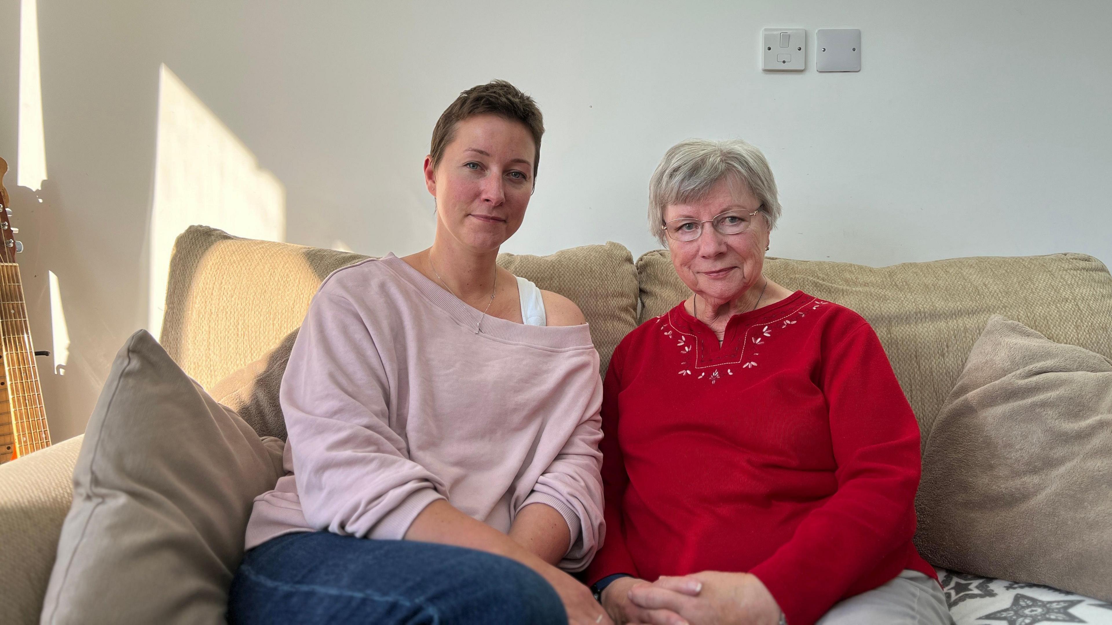 Margi and Ginny sit on a grey sofa looking at the came as they sit next to each other. Margi is wearing a pink jumper with her right shoulder showing and blue jeans. Ginny is wearing a red jumper with grey trousers and has glasses on. There's a guitar to the left of the sofa.