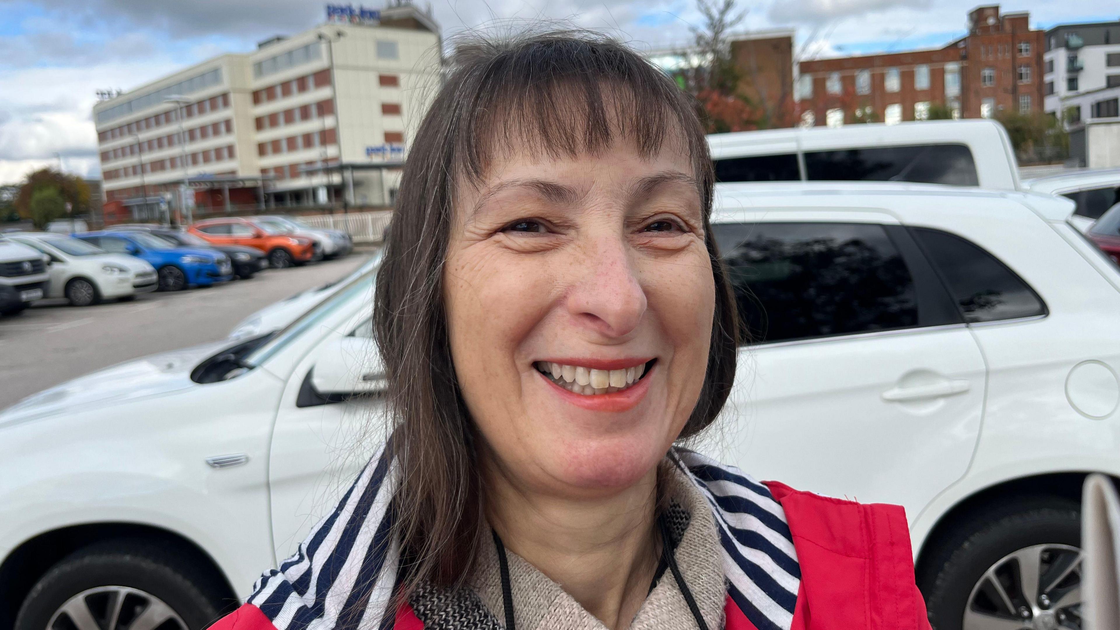 Barbara Patterson smiling for camera with a white car parked behind her. She has long black hair and is wearing a pink jacket with a navy and white striped hood.