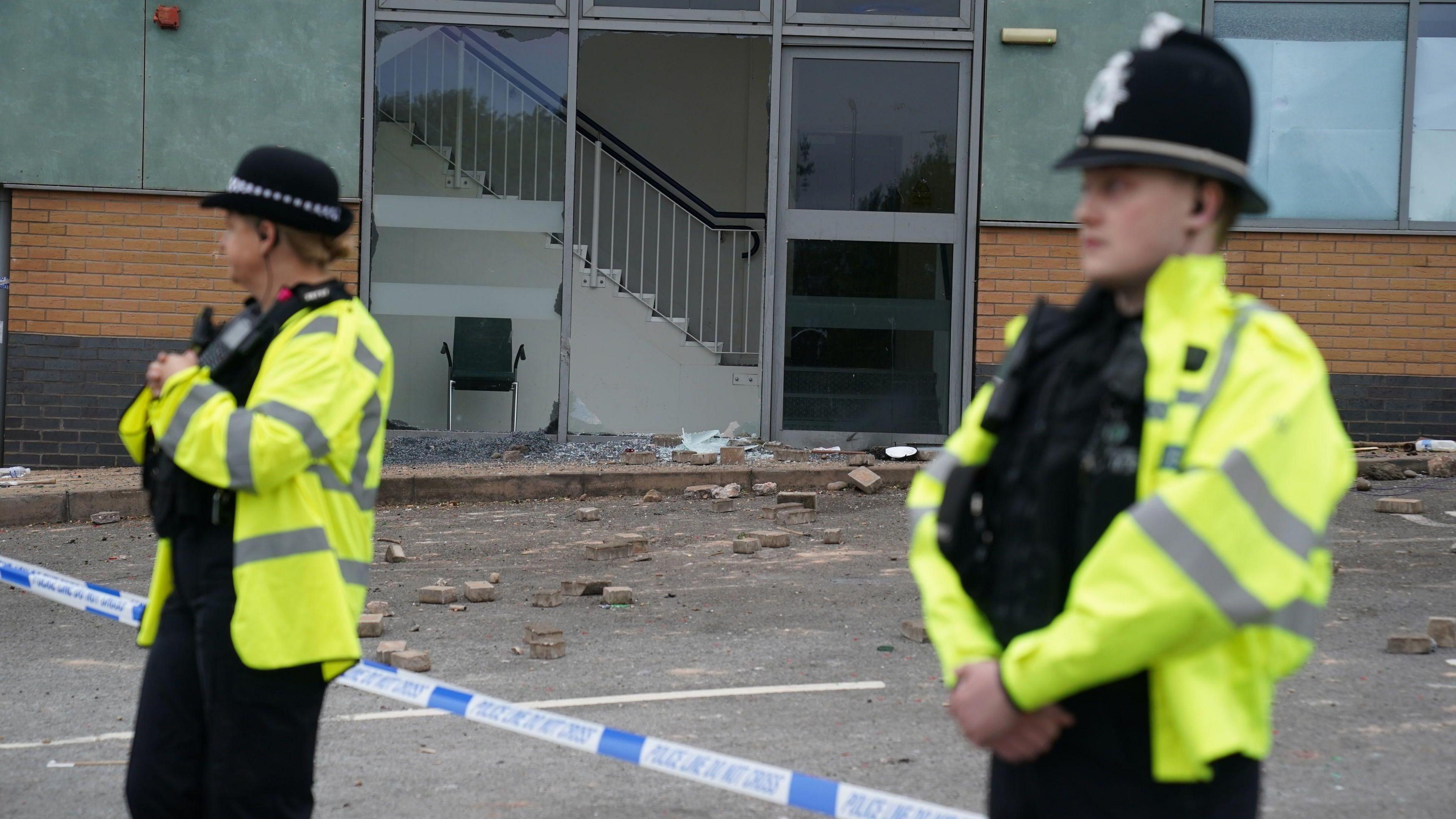 Police officers at the Holiday Inn Express in Tamworth, Staffordshire. They are wearing uniform and stood in front of blue police tape. In the background we can see part of the building has been smashed up with rocks and glass on the floor. 