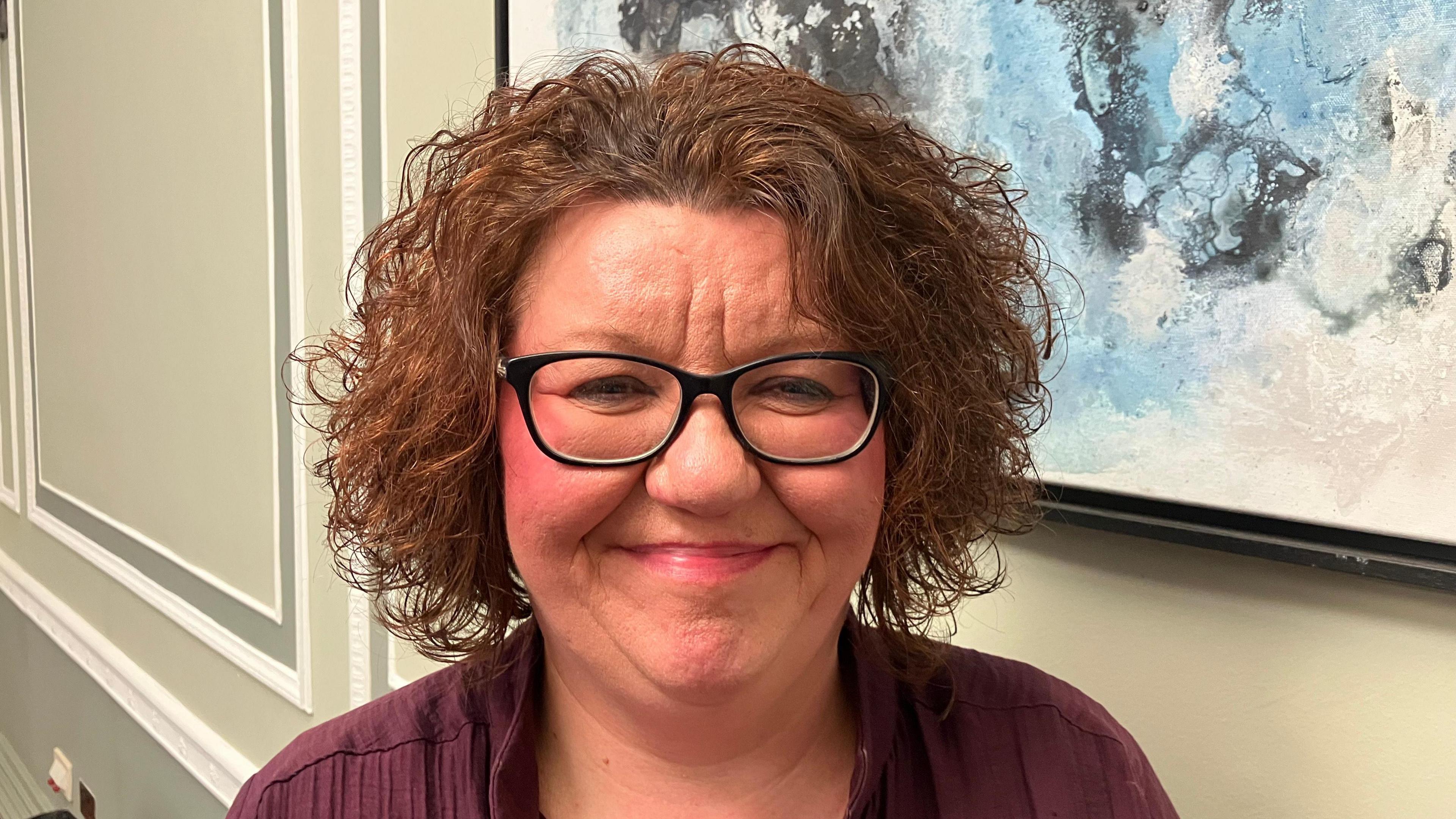 Julie Graham has short brown curly hair and is wearing makeup, black glasses and a purple top.