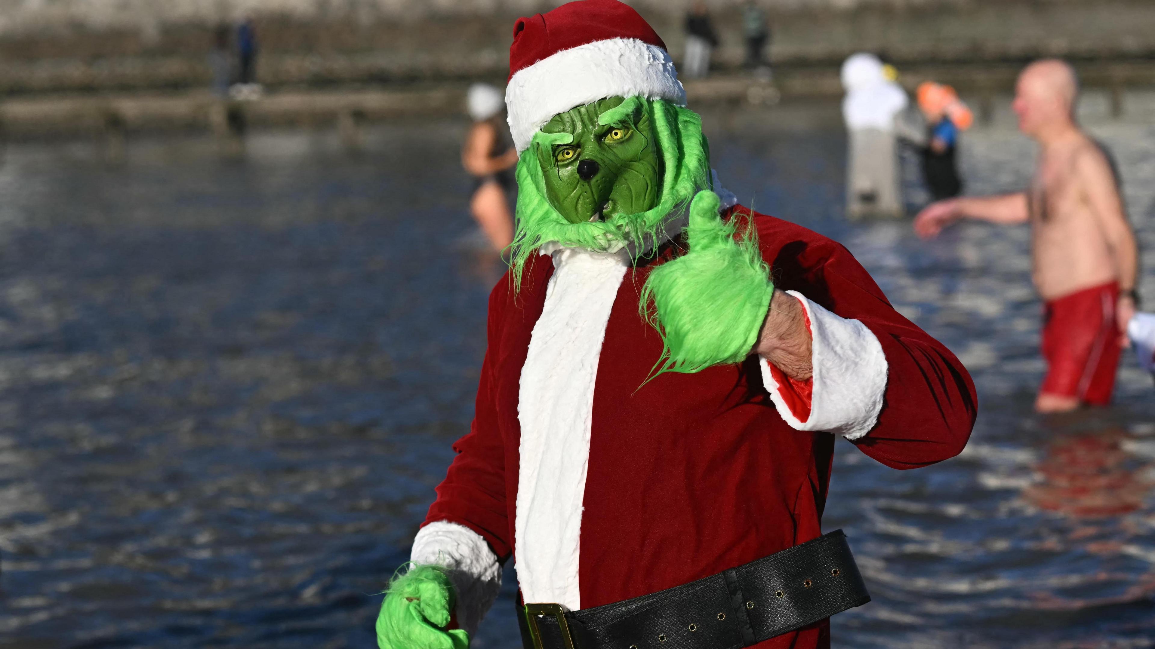 A person dressed up as the Grinch wearing a father Christmas suit, they put their thumb up at the camera. They are submerged to their waist in the sea.