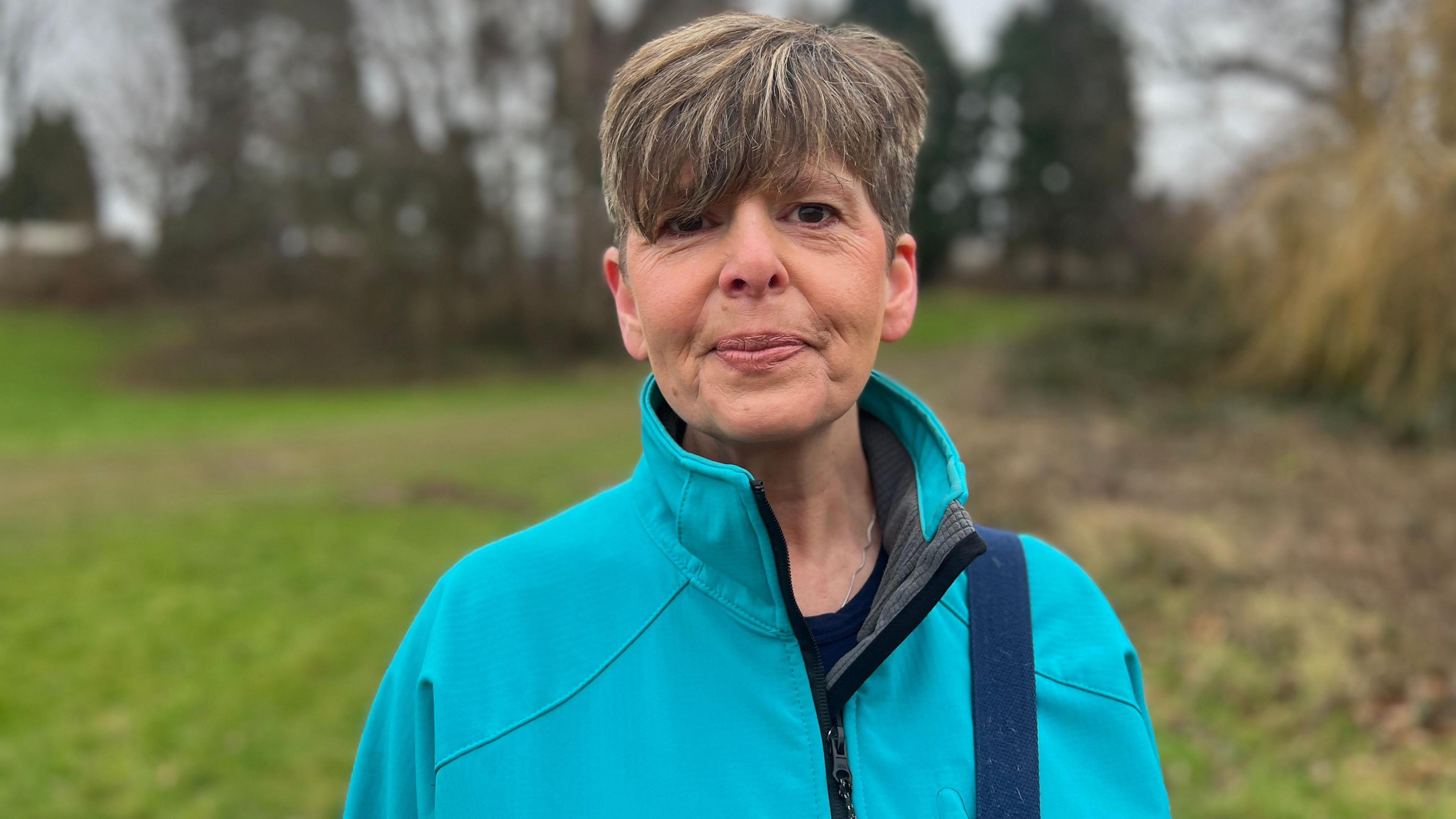 A woman who has short hair and a fringe that rests just above her eyes looks towards the camera. She is wearing a blue/turquoise jacket and is standing in what appears to be a park or a field