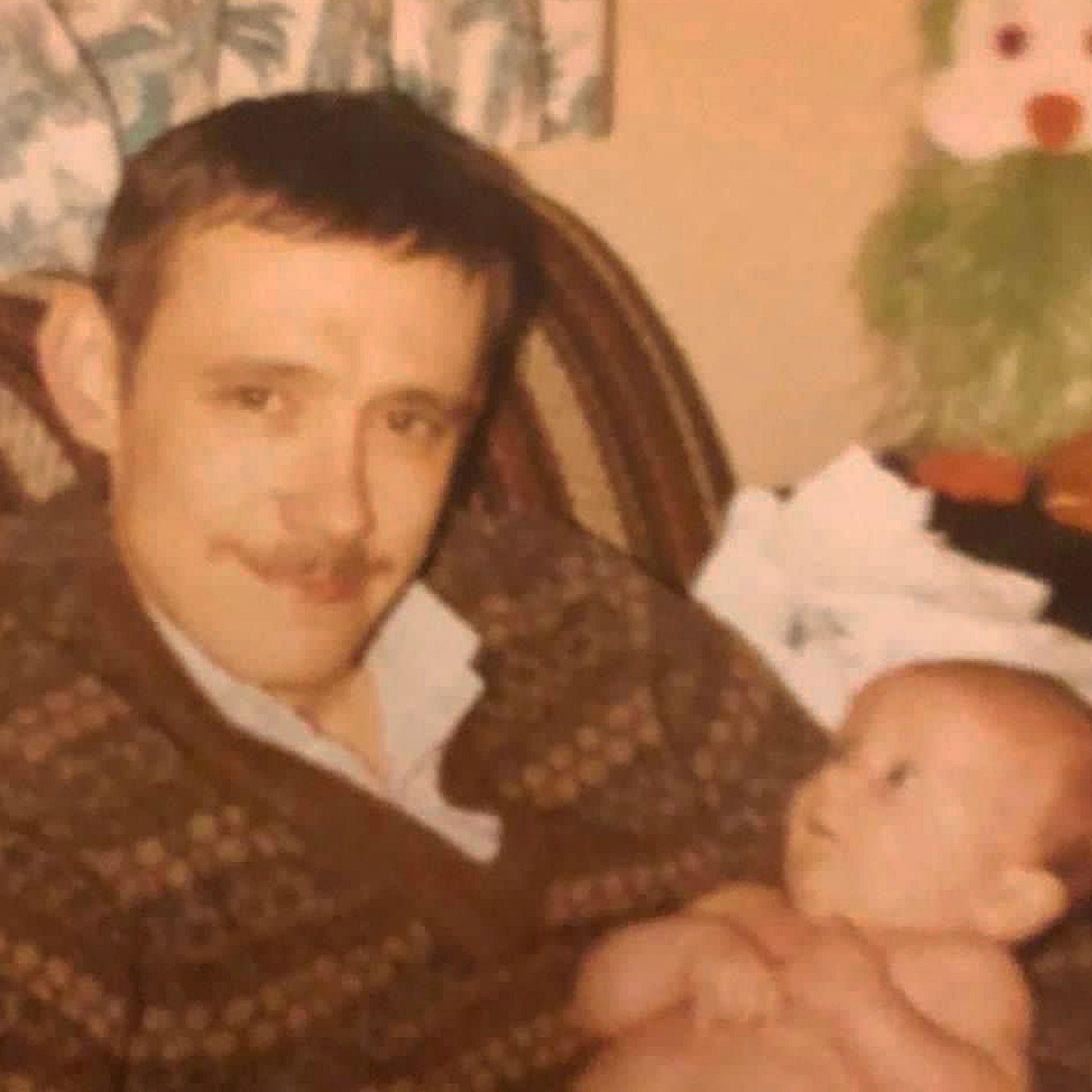 Ean Coutts when he was younger sitting on a seat while holding a baby. He has short hair and a moustache and is wearing a patterned brown, v-necked jumper with a shirt underneath.