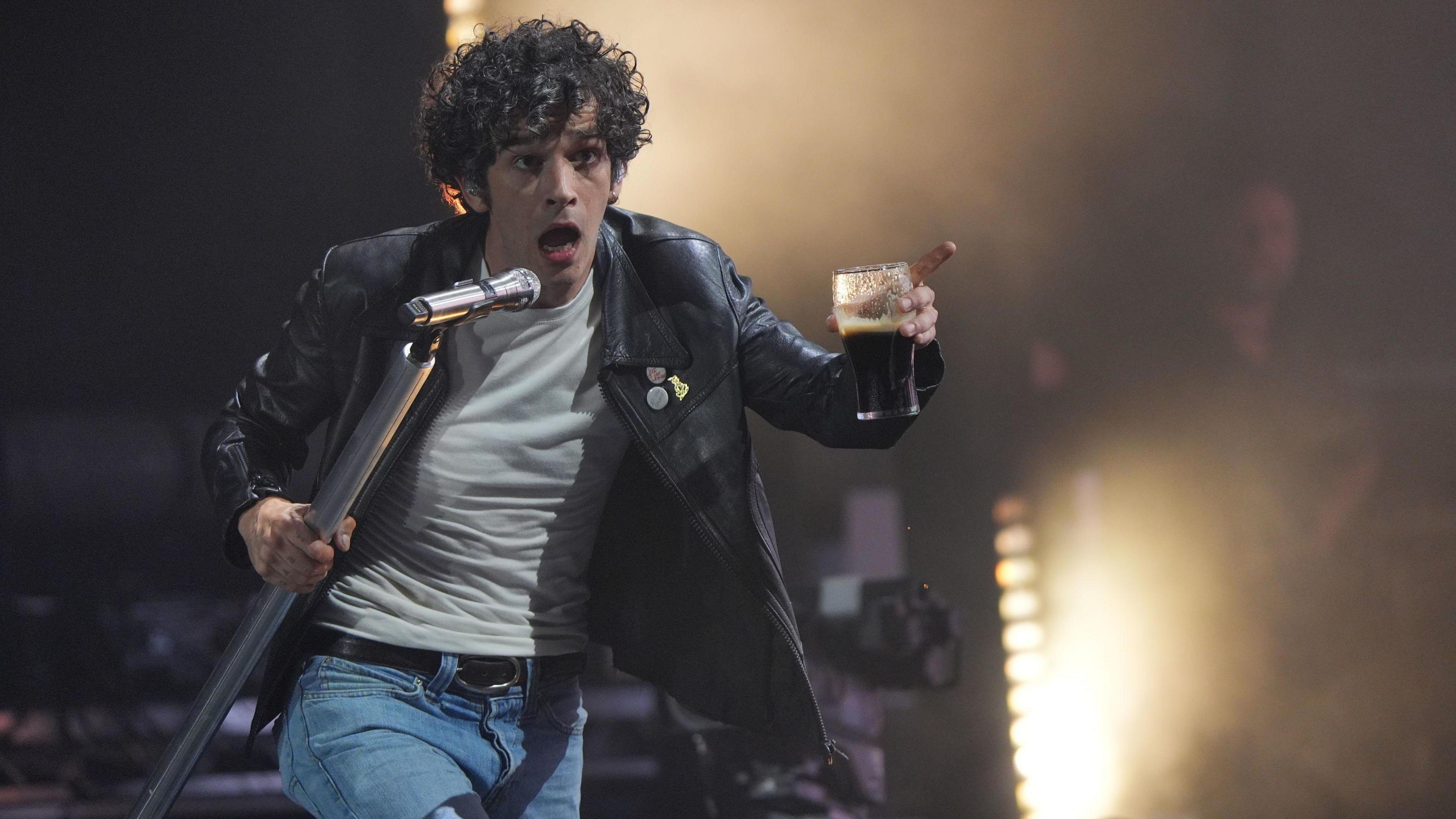 Matty Healey is on stage wearing a black leather jacket. He is walking towards the camera holding a microphone and a glass in each hand. He has black, curly hair.