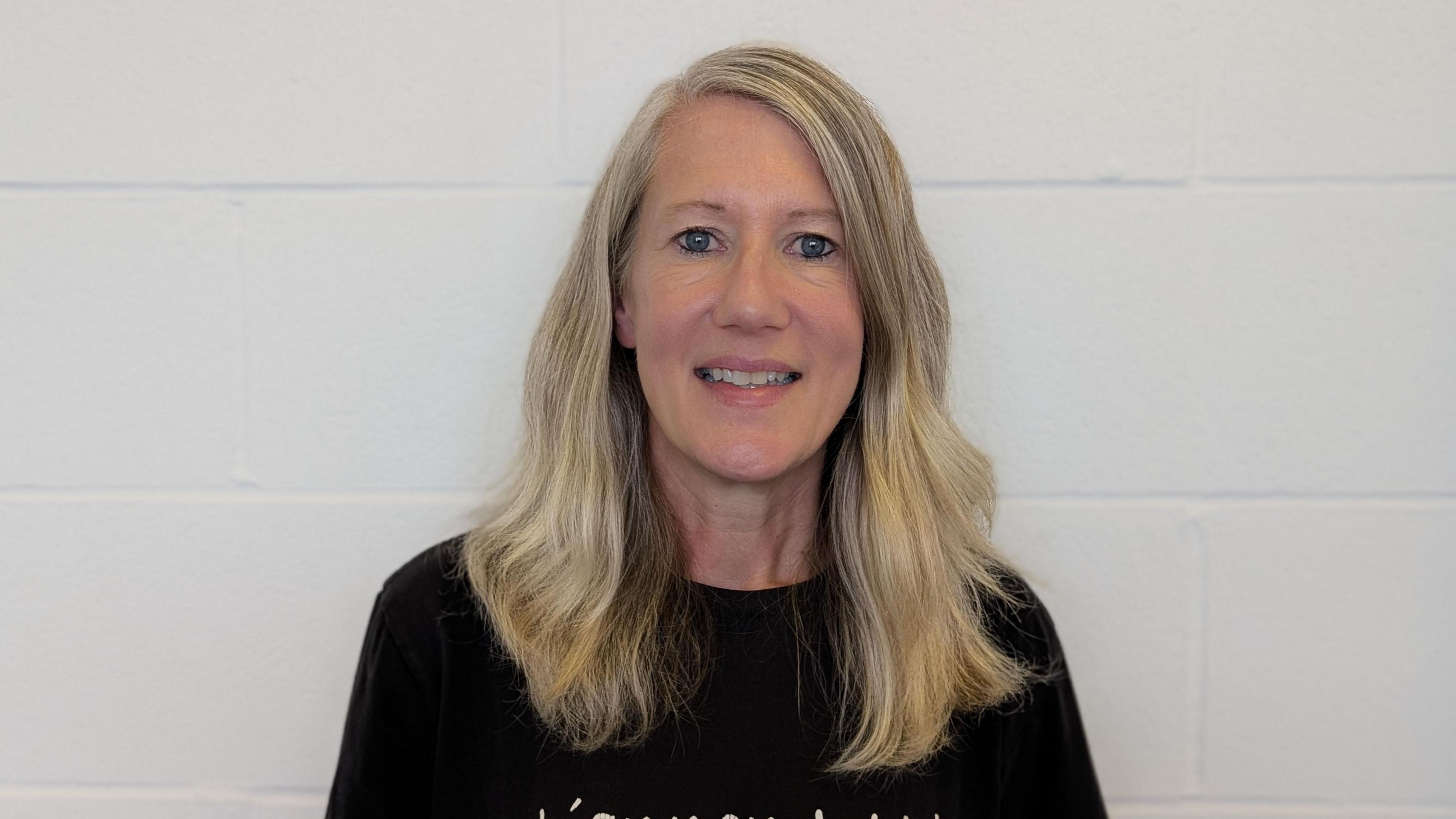 Rhiannon has long blonde hair and wears a black t-shirt. She's smiling at the camera against a white brick wall.