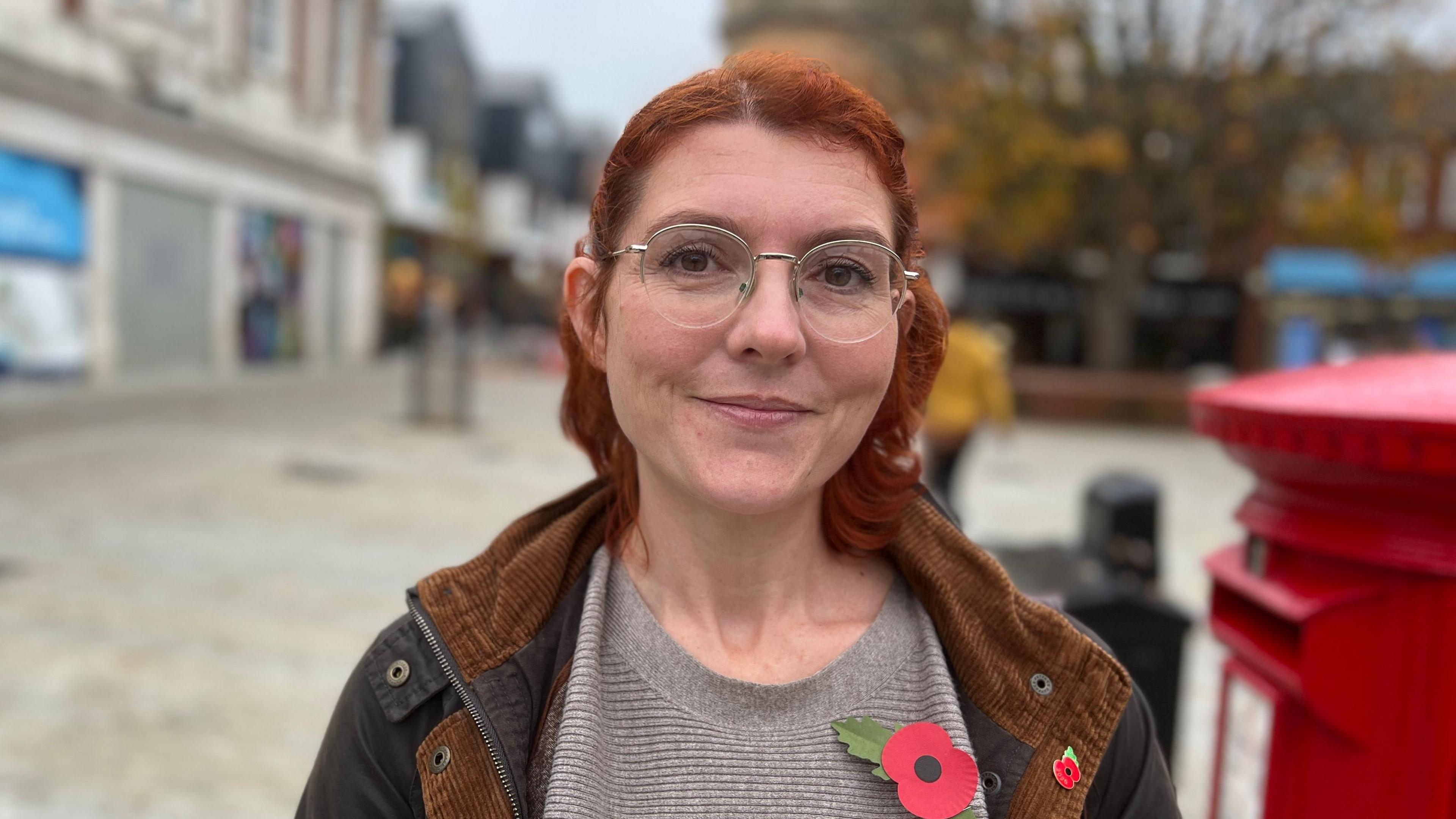 Natasha Osben stands in a town centre which is blurred behind her. She smiles at the camera. She has short red hair and wears round glasses, a black and brown coat with a grey top underneath. She has a large poppy pinned to her top and another smaller poppy pinned to her coat. A red post box can be seen to the right of her. 