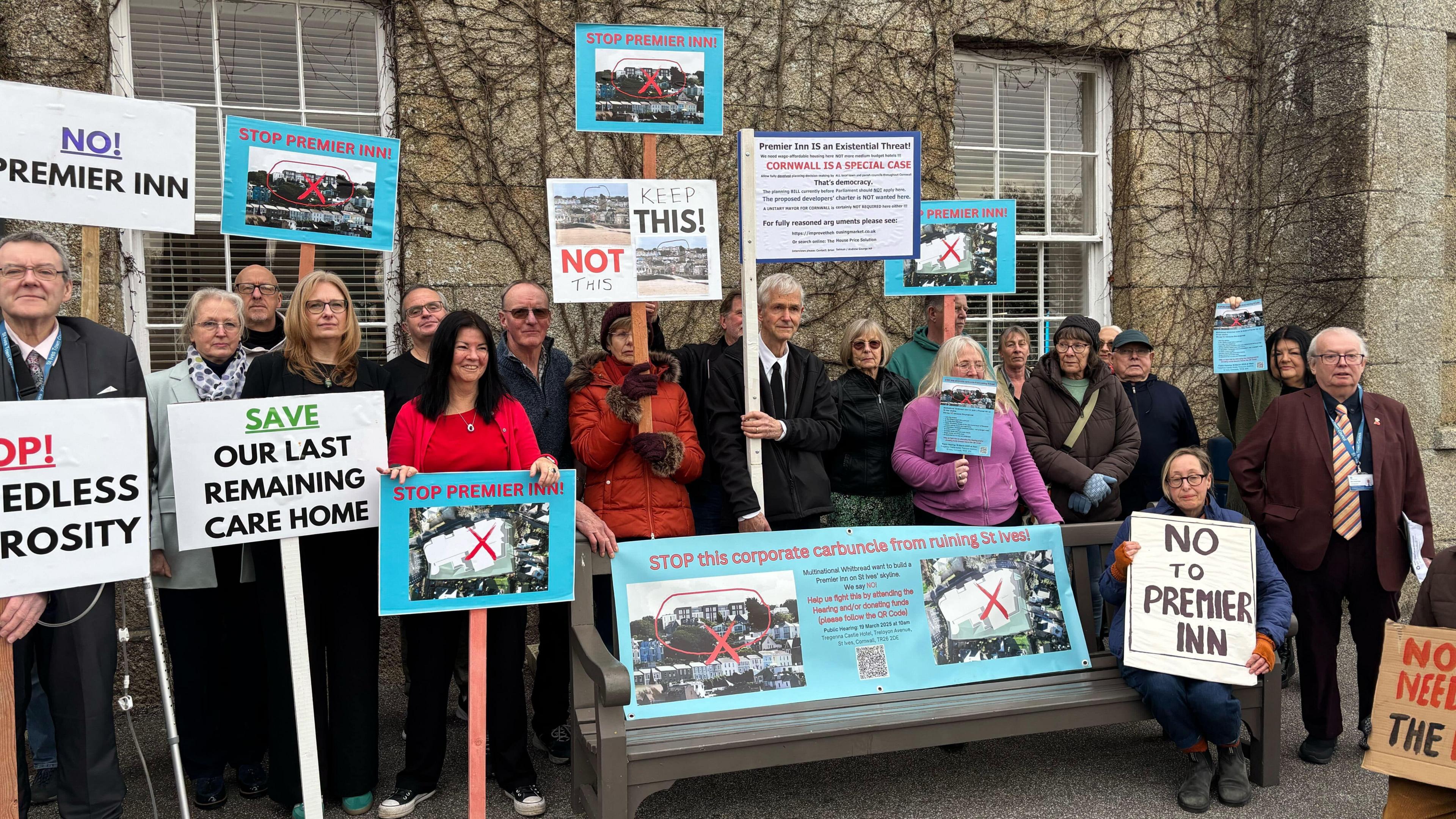 About 20 people outside a hotel holding placards and banners against the proposed plans. There is a printed banner attached to a bench with a lady sat next to it with a banner saying 'No to Premier Inn'.