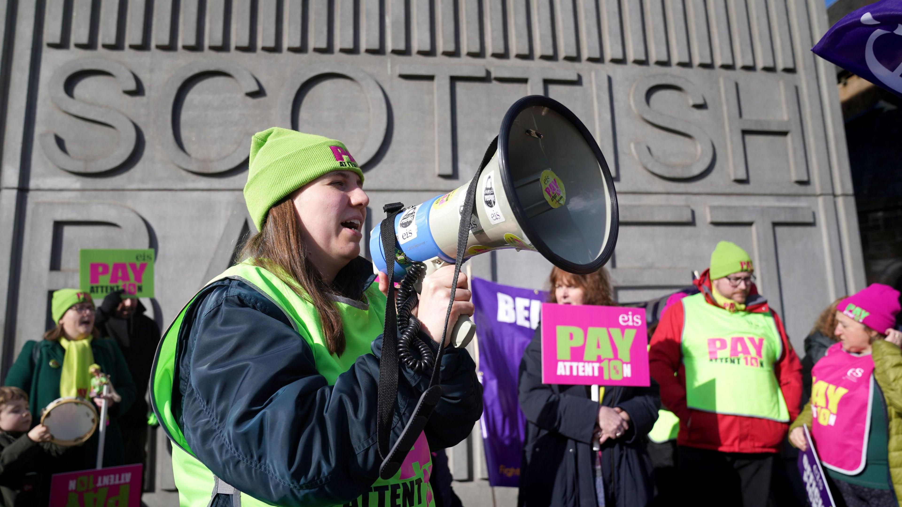 Picture of a school strike from 2024, with teachers wearing colourful EIS branded tabards and a woman speaking into a megaphone in the foreground