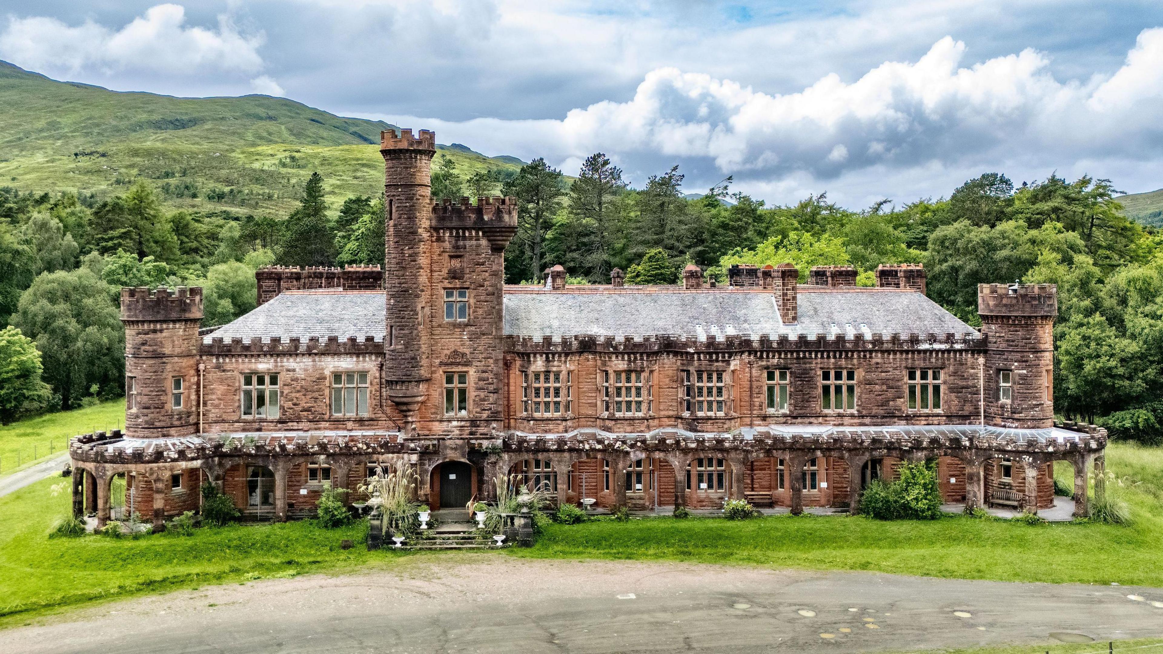 An exterior view of the red sandstone lodge with its ornate turrets and battlement-like rooftops. The lodge is set in an grounds with large areas of grass and trees.