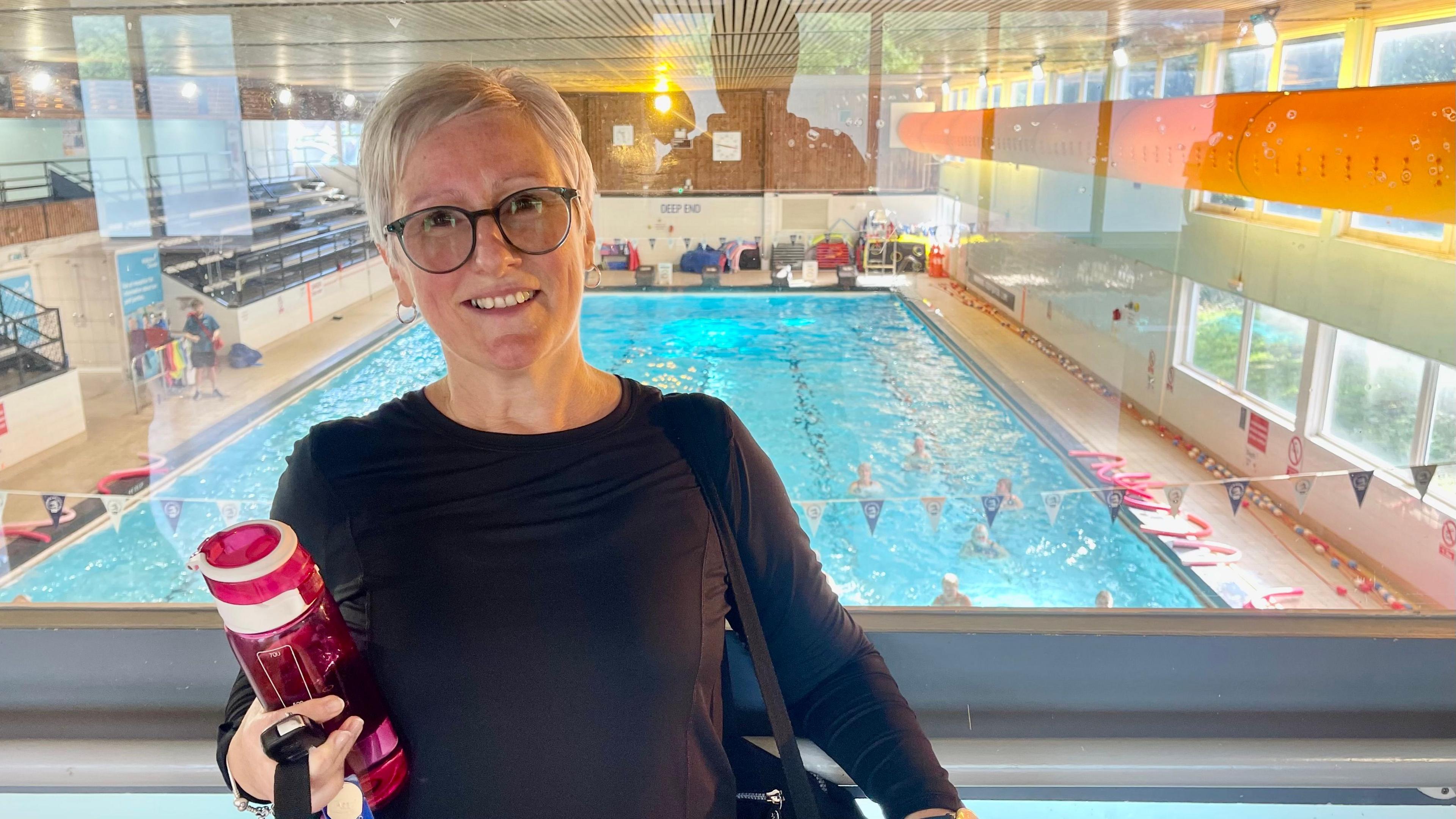 A woman will dark round glasses, short blonde hair, a black long sleeved gym top, a black sports bag and a pink water bottle is stood on the glass balcony of a swimming pool. Seven swimmers can be seen at one end of the pool. There are floats, seating areas and a lifeguard in a blue t-shirt and short beside the pool.