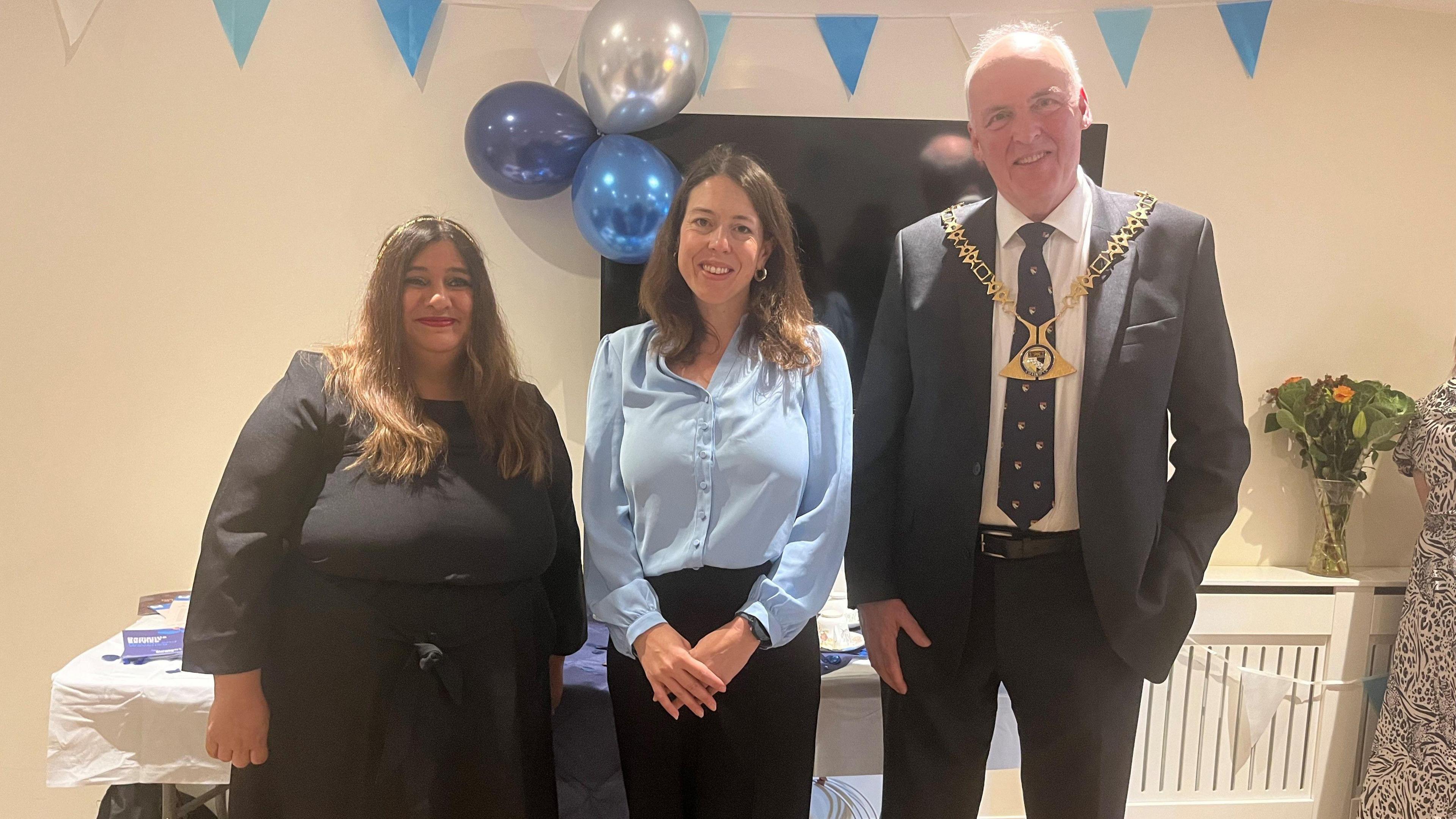 Three people are standing side by side in front of a wall with blue balloons and bunting on. The two people on the left are women and the other person is a man in a suit 
