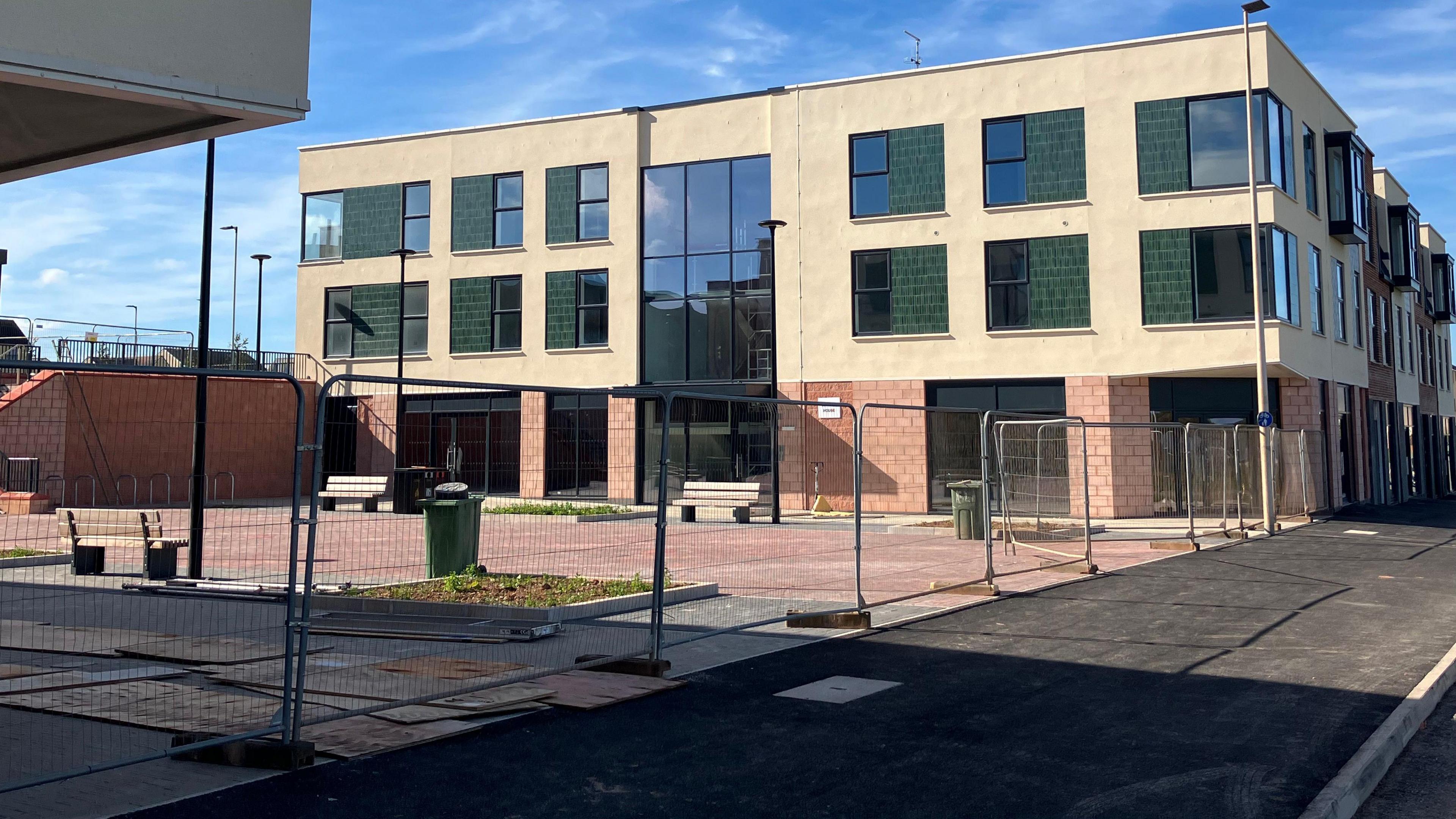 A red brick and light brown new build with 12 windows at the front on the second and first floor. There is fencing surrounding the new building with a black tarmac pathway on the other side. There is also a green wheelie bin to the left inside of the fencing with a red brick wall 