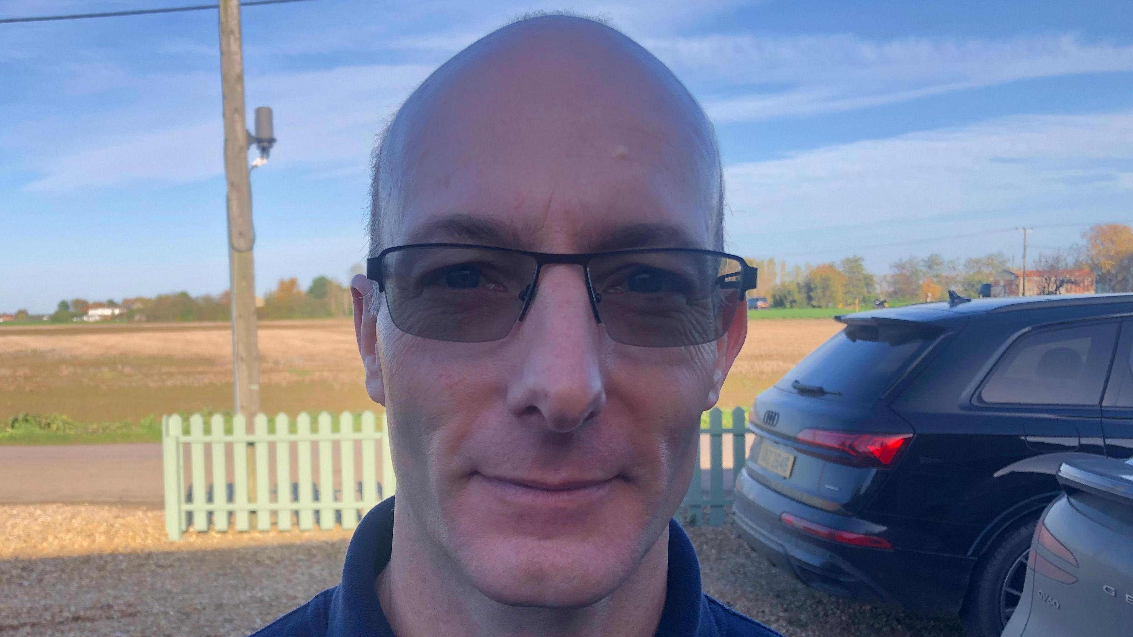 Andrew Baldwin, 52 has a slim face, with balding head, tinted glasses and a blue fleece top. Behind him are farmers fields with light brown ploughed soil, there are two cars parked on the right and a light green picket fence beyond them across a gravel driveway.