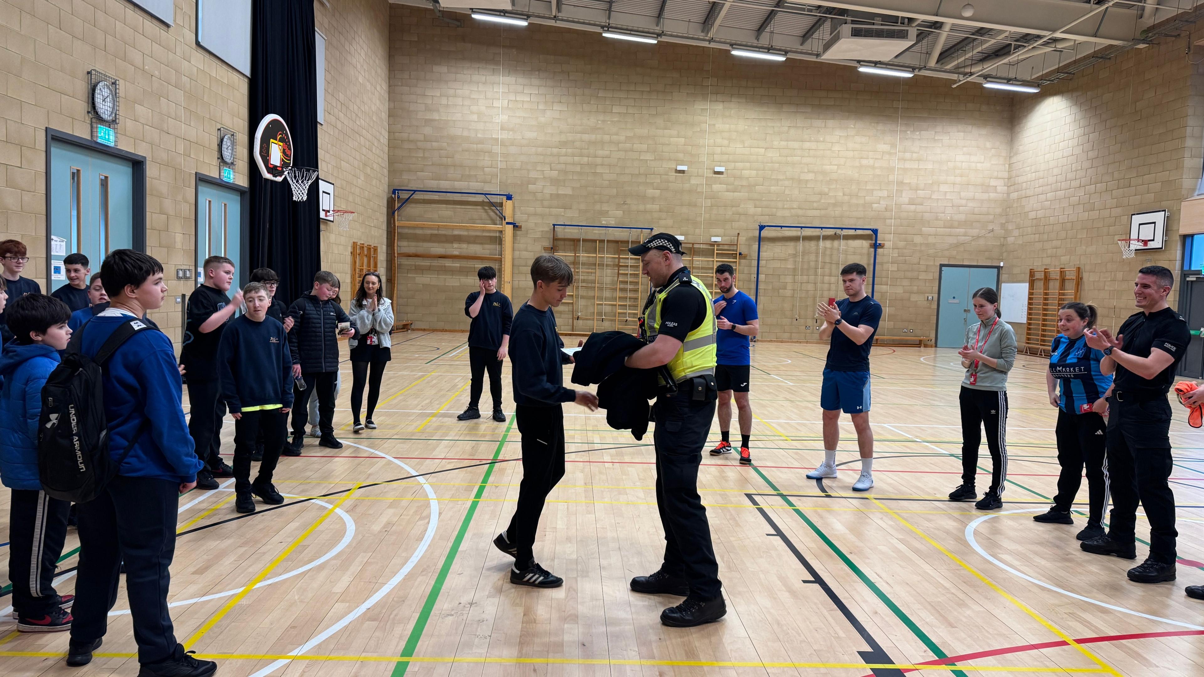 Fear de chluicheadairean Sgoil MhicNeacail a' togail duais airson cluicheadair na seachdaine agus e a' gabhail grèim air làmh phoilis a tha ga thoirt seachad. 