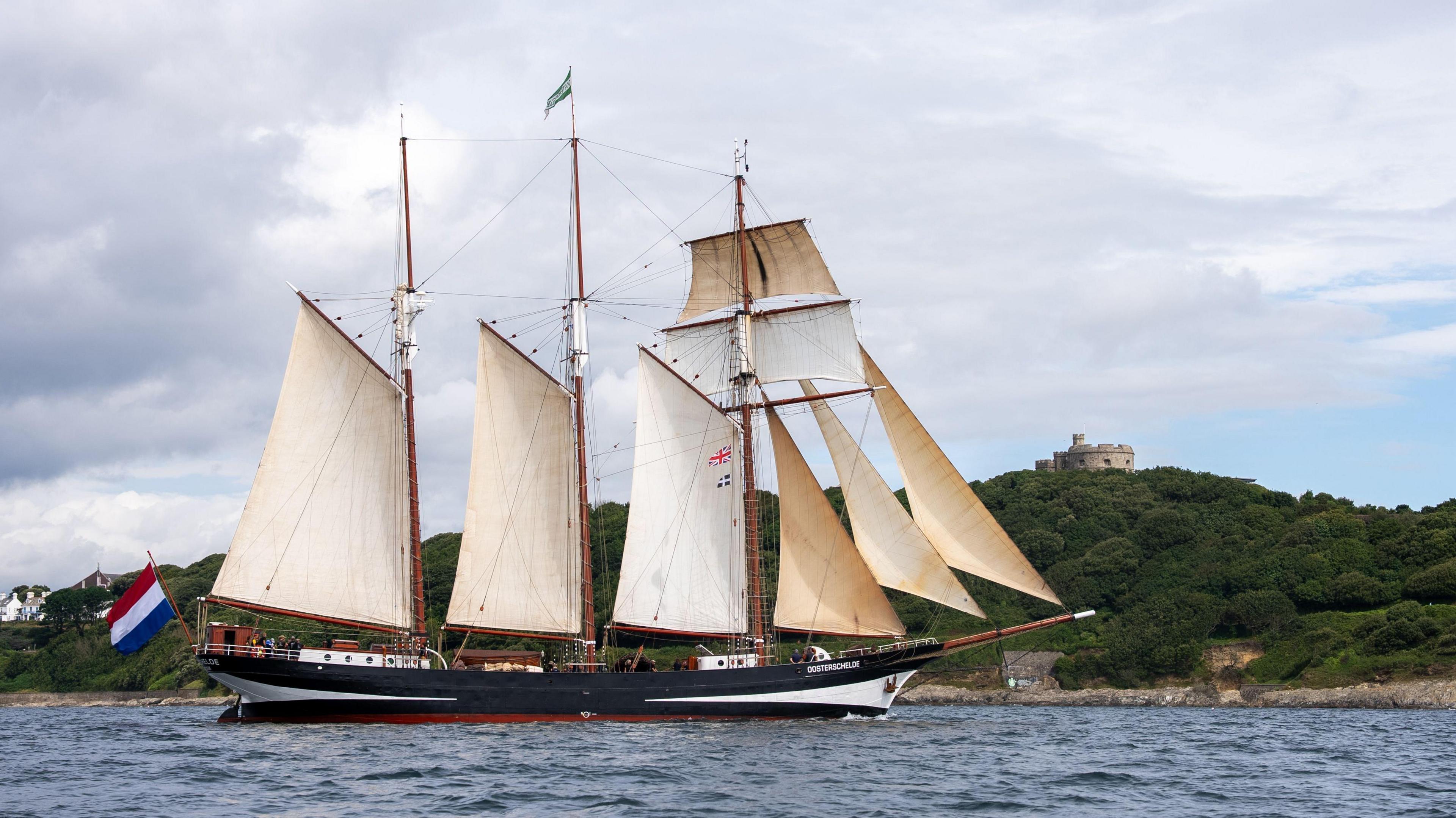 The Oosterschelde taken from the side. It is an historic tall ship with eight sails. In the background is a green hill with a small fort on top. A Dutch flag hangs off the back of the ship. 