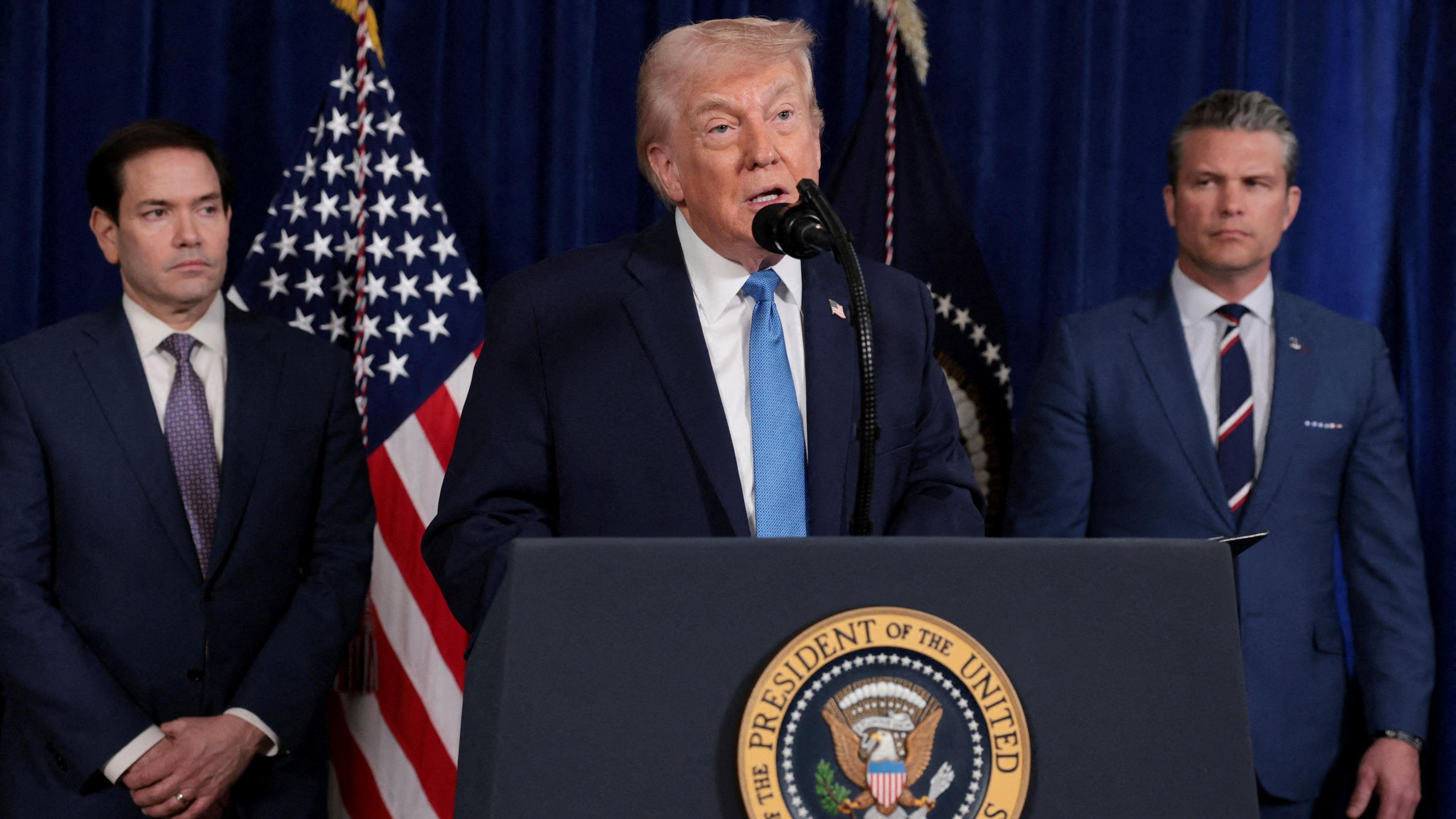 President Donald Trump makes a speech with a US flag behind him. Stood next to him is Secretary of State Marco Rubio and Secretary of Defense Pete Hegseth, who are looking forwards during the press conference.