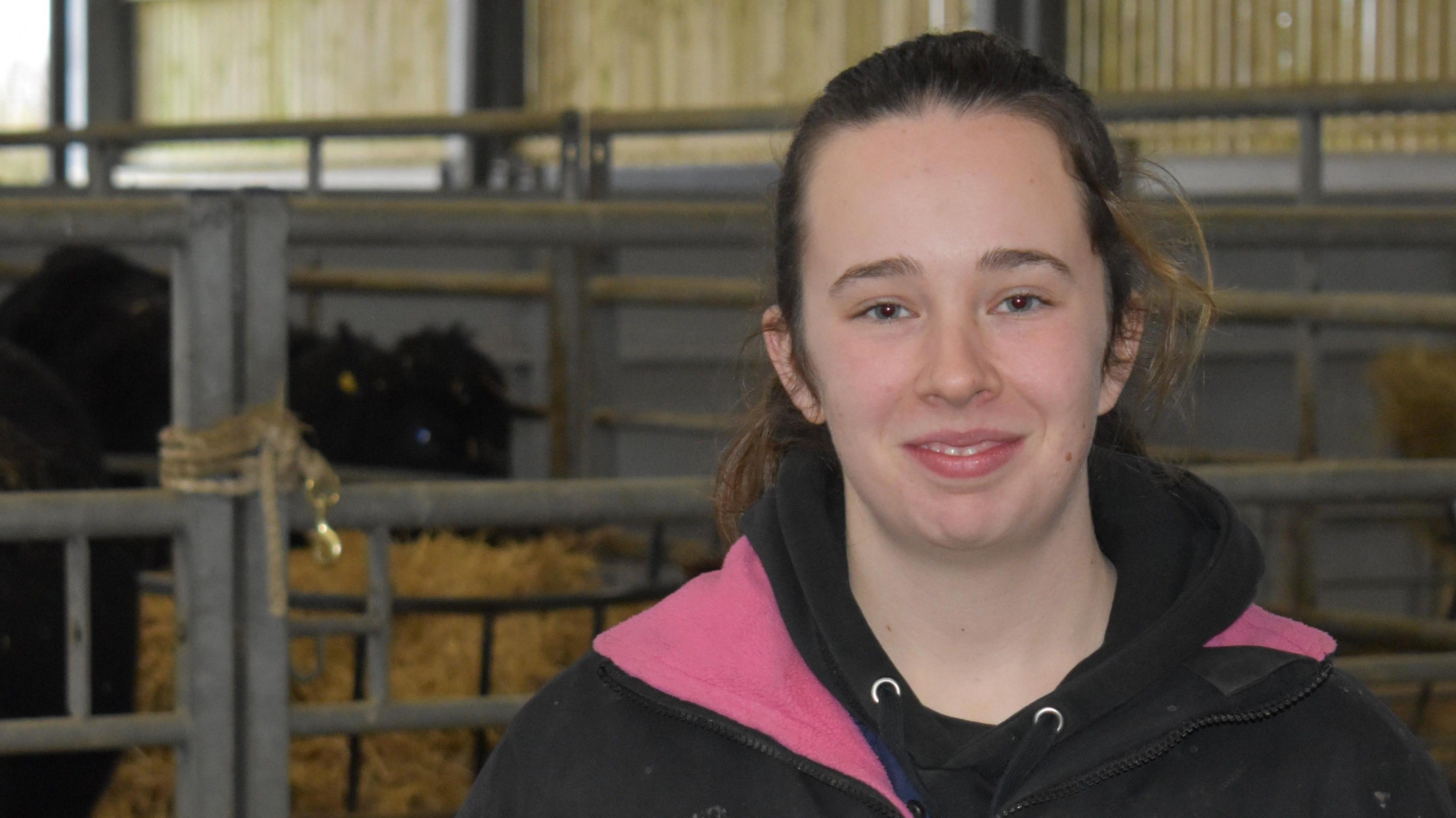 Sophia is standing in a barn. She is wearing a dark coat with pink lining visible. Her brown hair is tied back. Behind her, black cattle can be seen standing in their pen