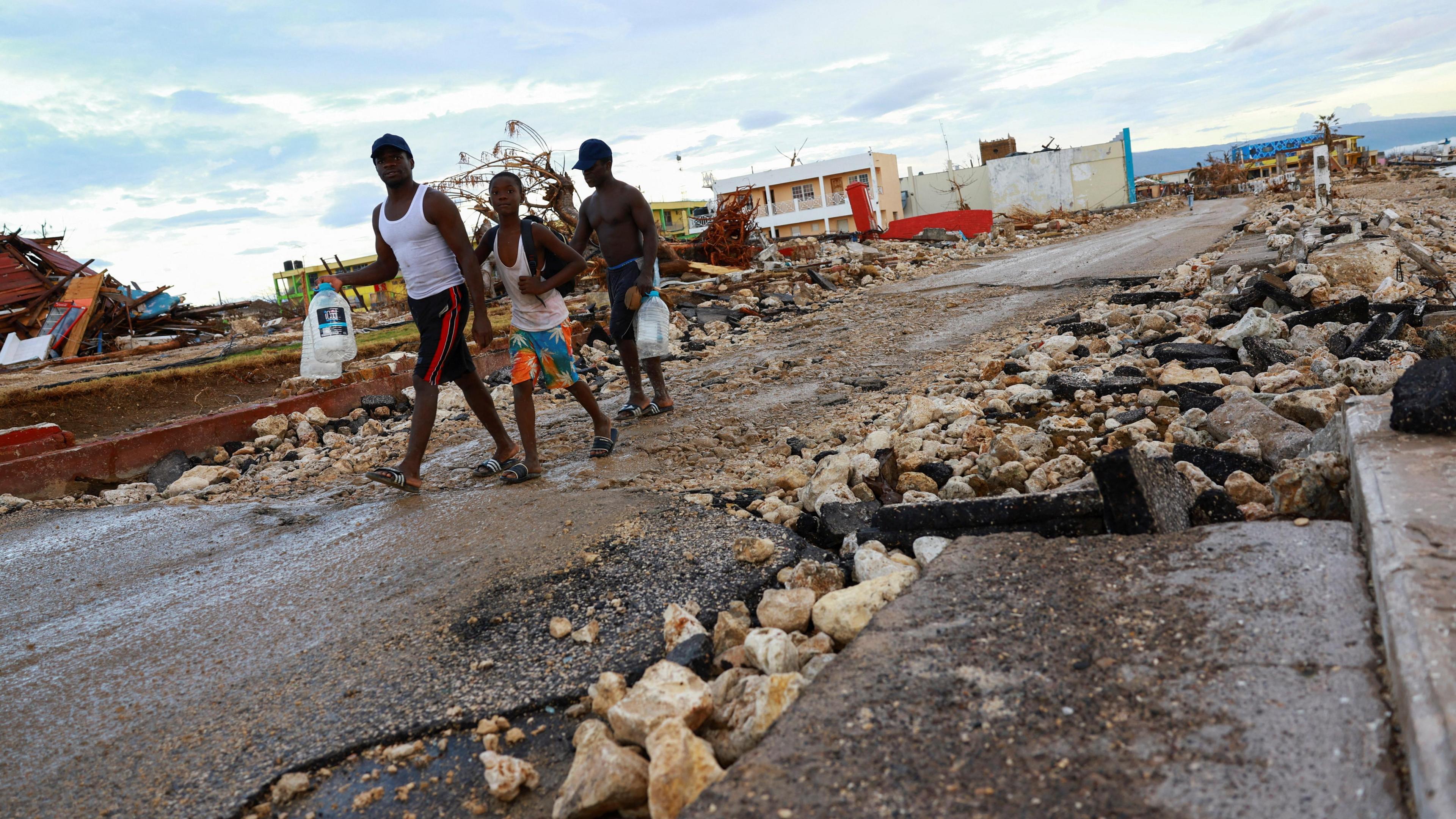 Three men walk along a street surrounded by rubble and remnants of building - a scene of devastation as far as the camera can see. Two carry large bottles of water.