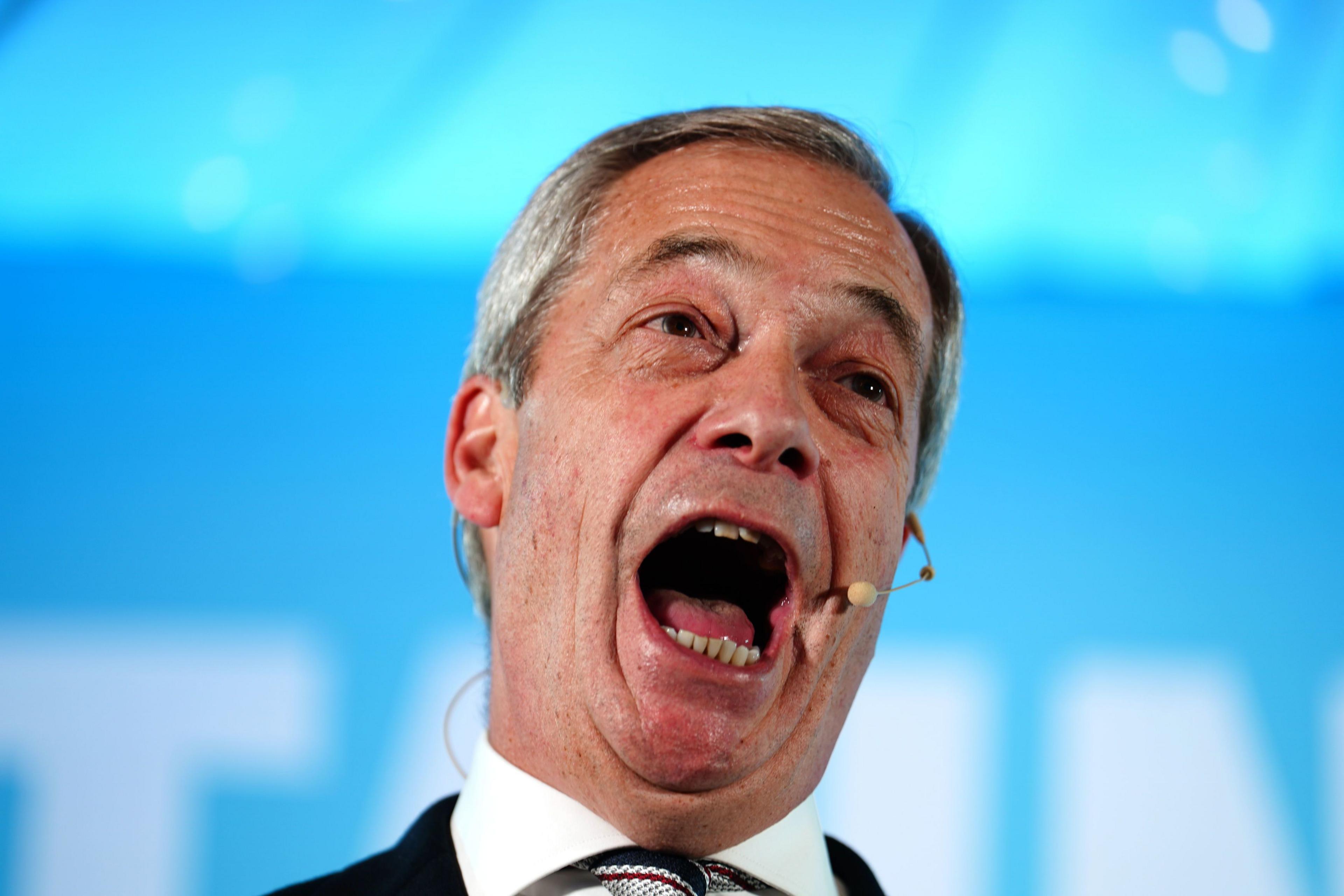 Close up of Farage speaking on stage. His mouth is wide open and he is wearing a nude coloured headset