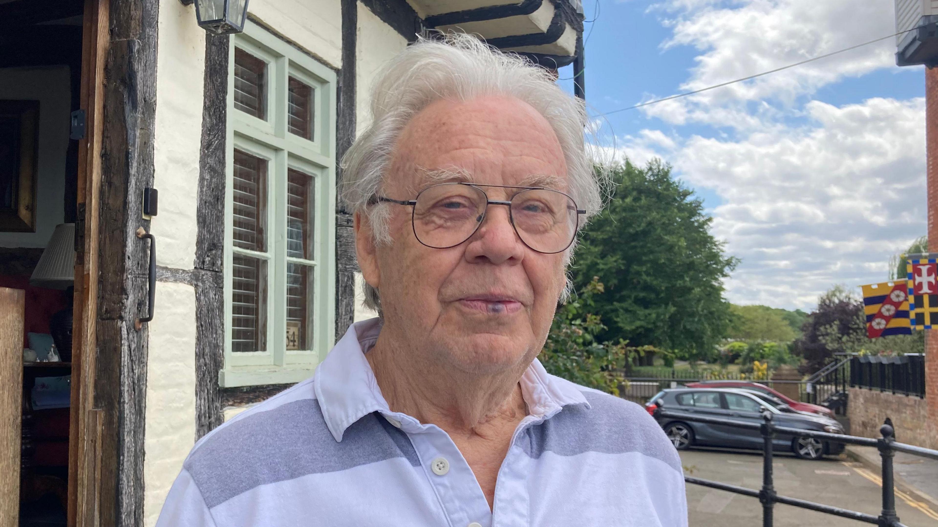 A photo of a main with white hair standing in Tewkesbury. He is wearing glasses and a grey and white shirt. He is stood in front of a building with brown timbers, white walls and a green window frame