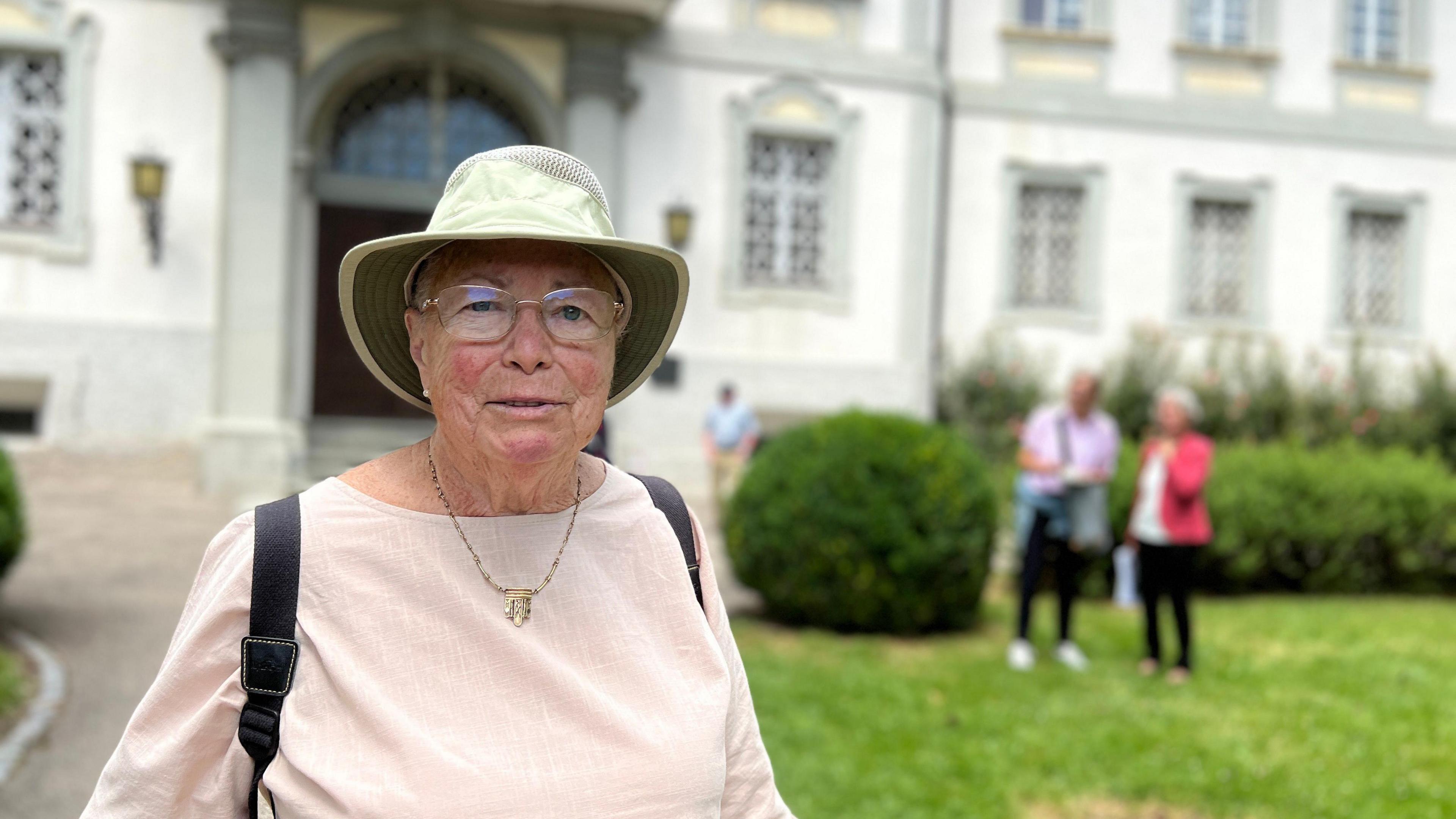 Angela is wearing a light pink blouse, green sun hat, and glasses stands in front of a large white building with multiple windows and an arched entrance. In the background, two people stand on the grass near bushes, and another person walks along a pathway.