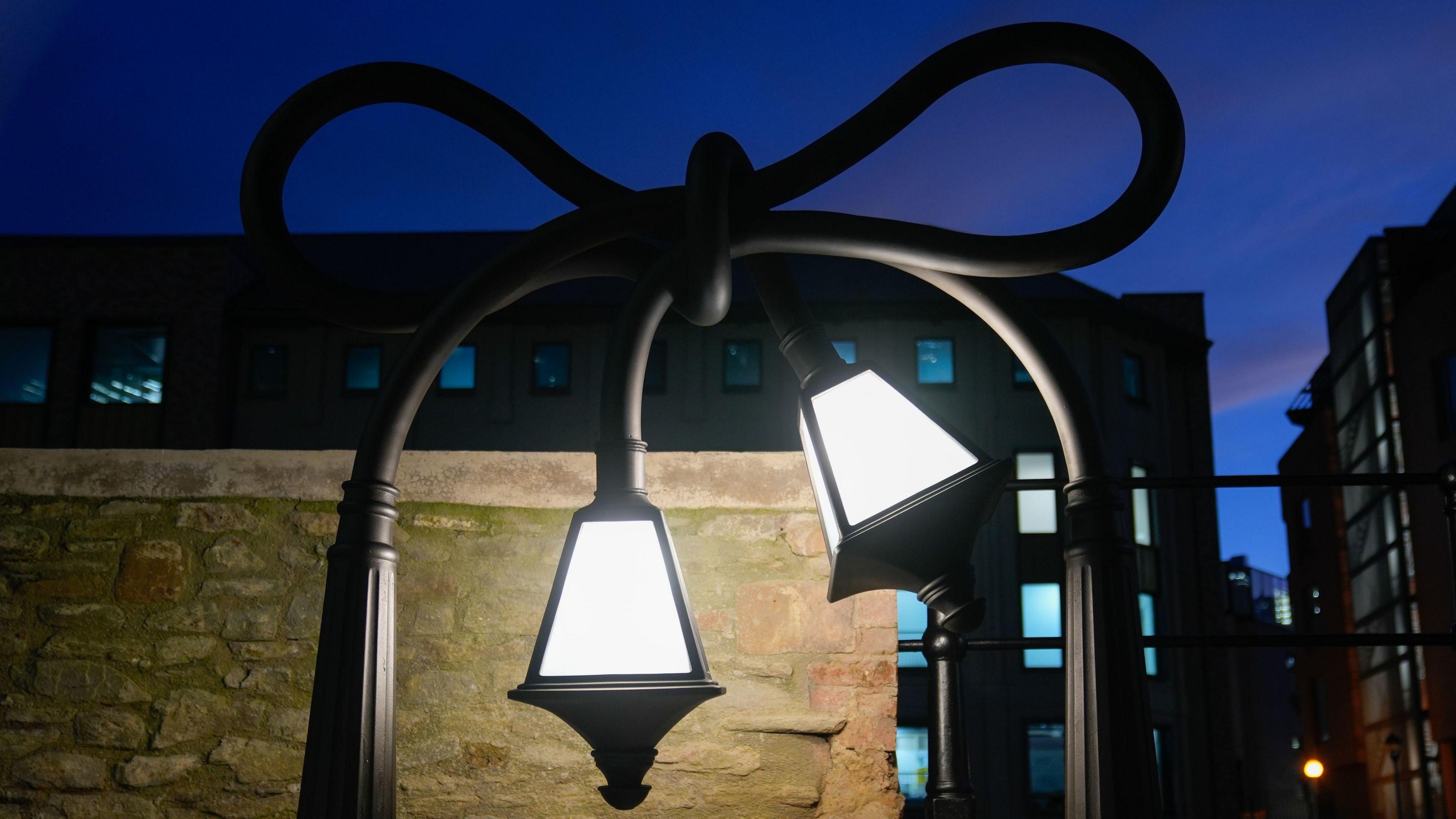 A view of an artwork of two old style lampposts tied in a knot on a public pathway by a canal. The picture is taken at night and the lamps are on. The artwork, in Bristol, was created by Alex Chinneck