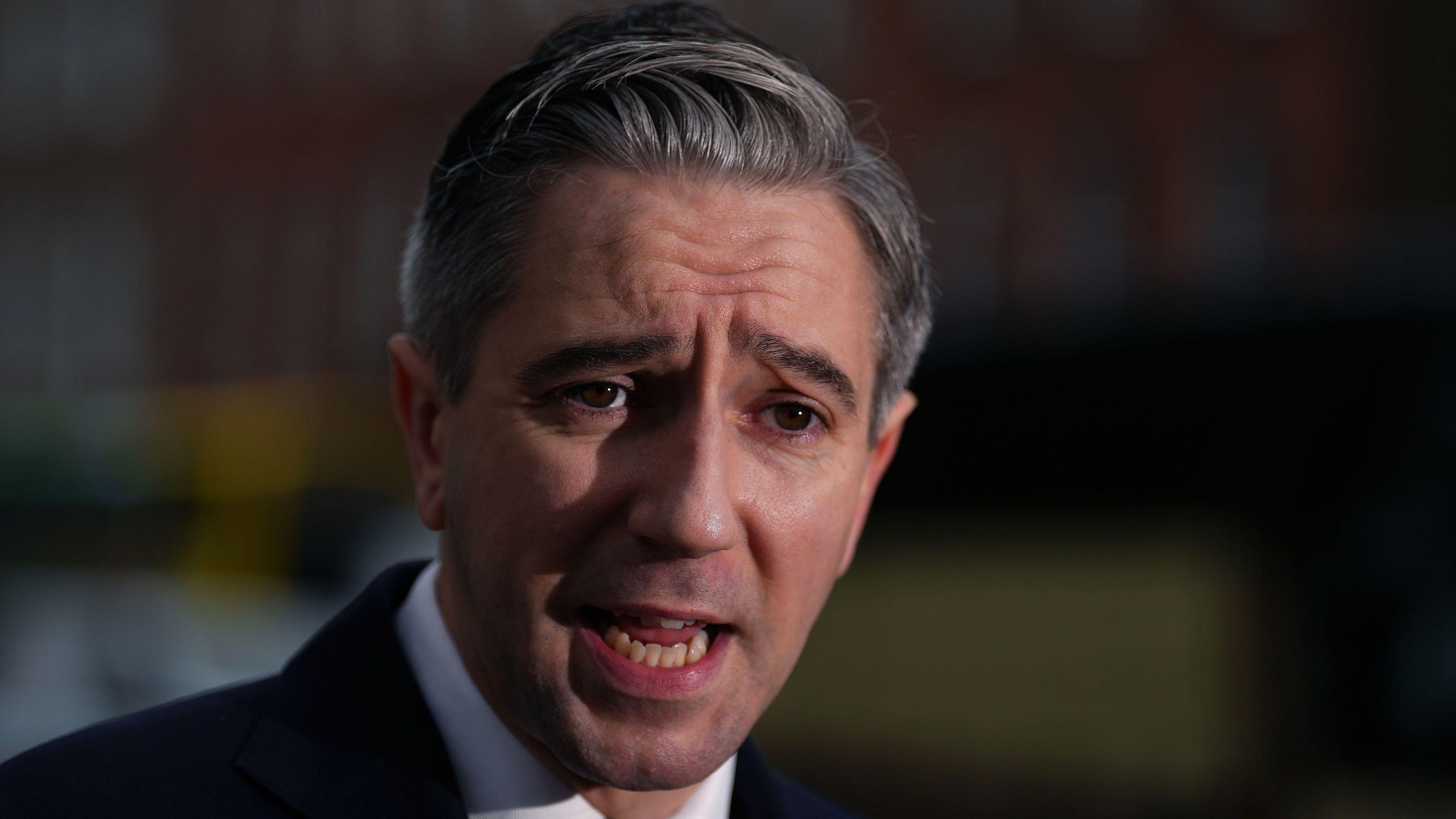 Simon Harris has short grey hair and is wearing a black suit jacket, white shirt and black tie in the picture.