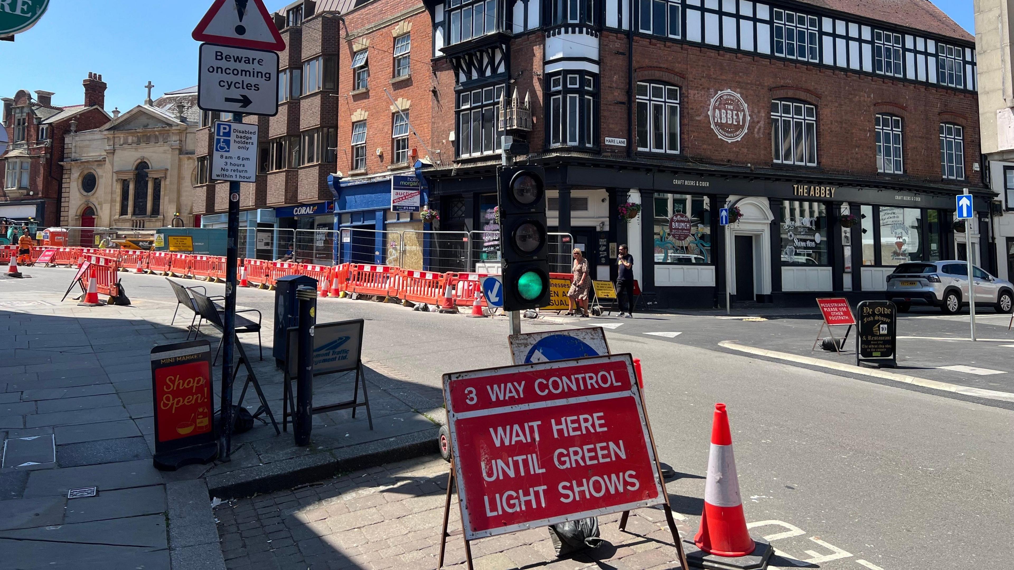 A traffic junction with a red metal sign with the words "3 way control. Wait here until green light shows" in the foreground. Orange barriers and fencing can be seen on the other side of the largely deserted road.