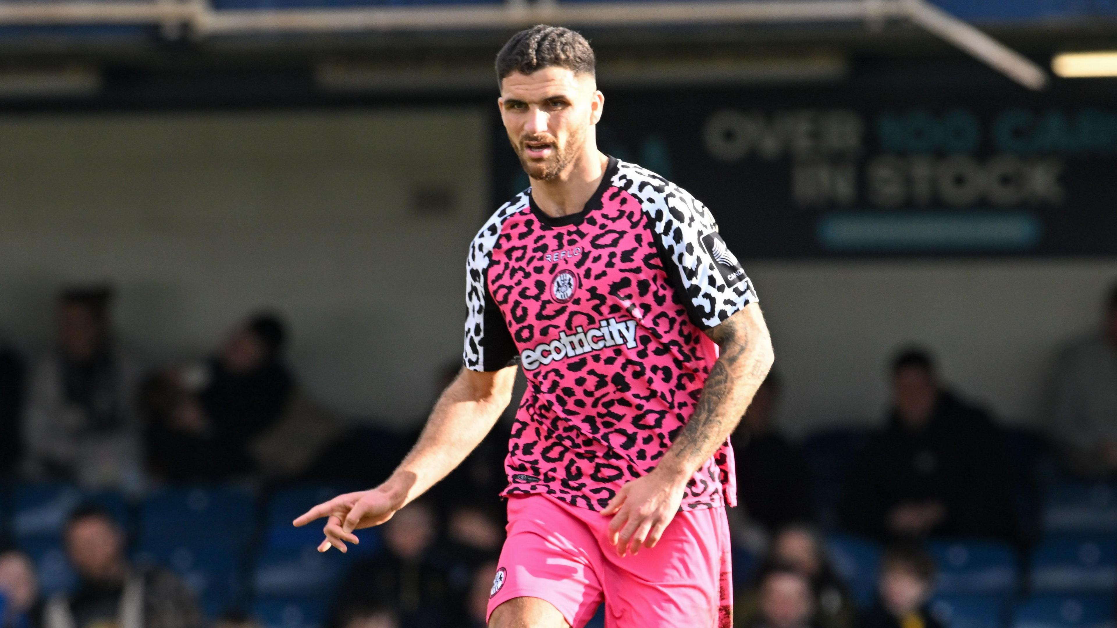 Ryan Inniss running on the pitch, wearing Forest Green's pink away kit, with his right arm out at his side and index finger extended