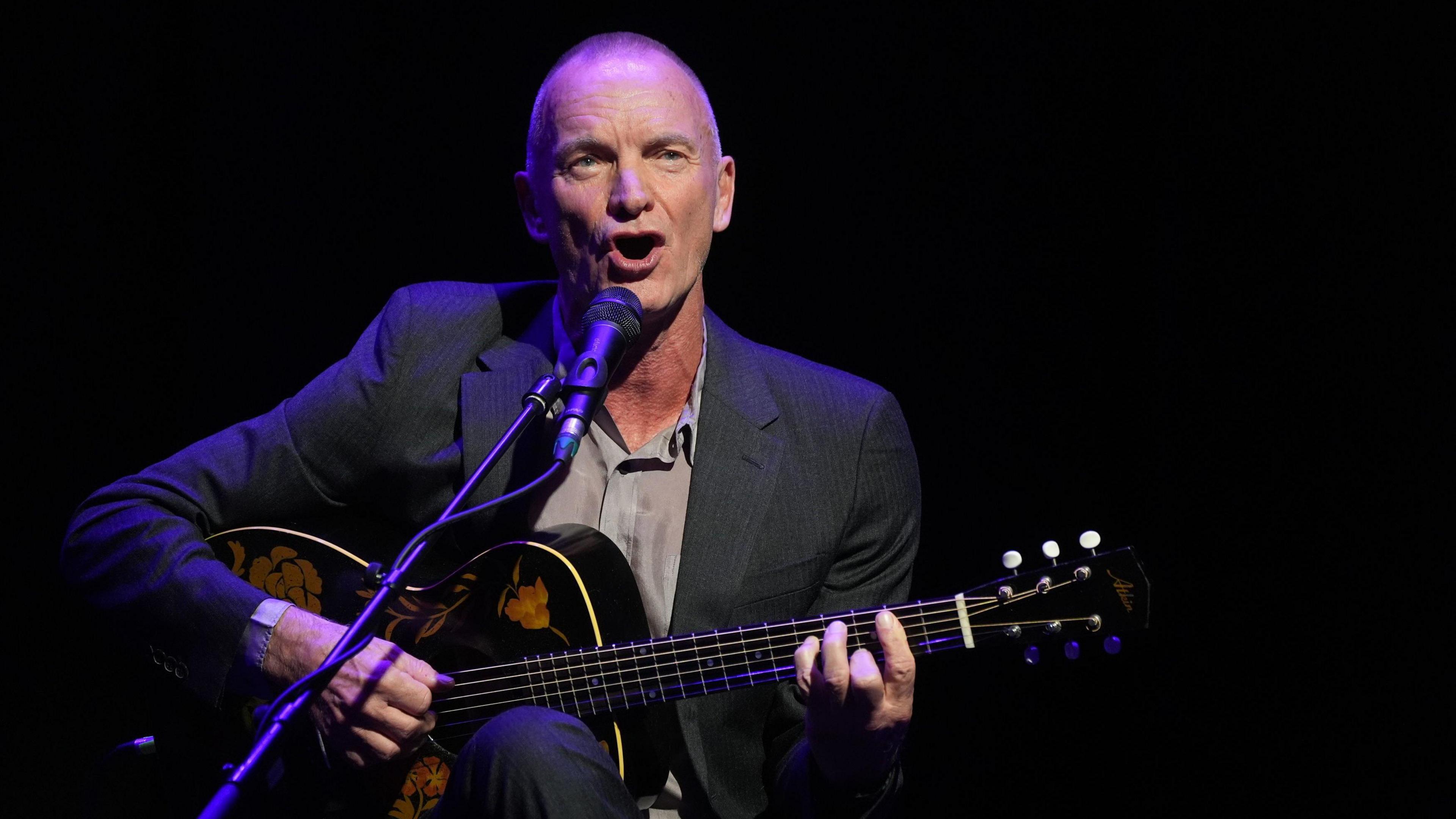 Sting performing at the Baltic Centre for Contemporary Art. He has a shaved head and is wearing a grey suit and shirt. He is sitting down as he sings and plays a guitar next to a microphone.