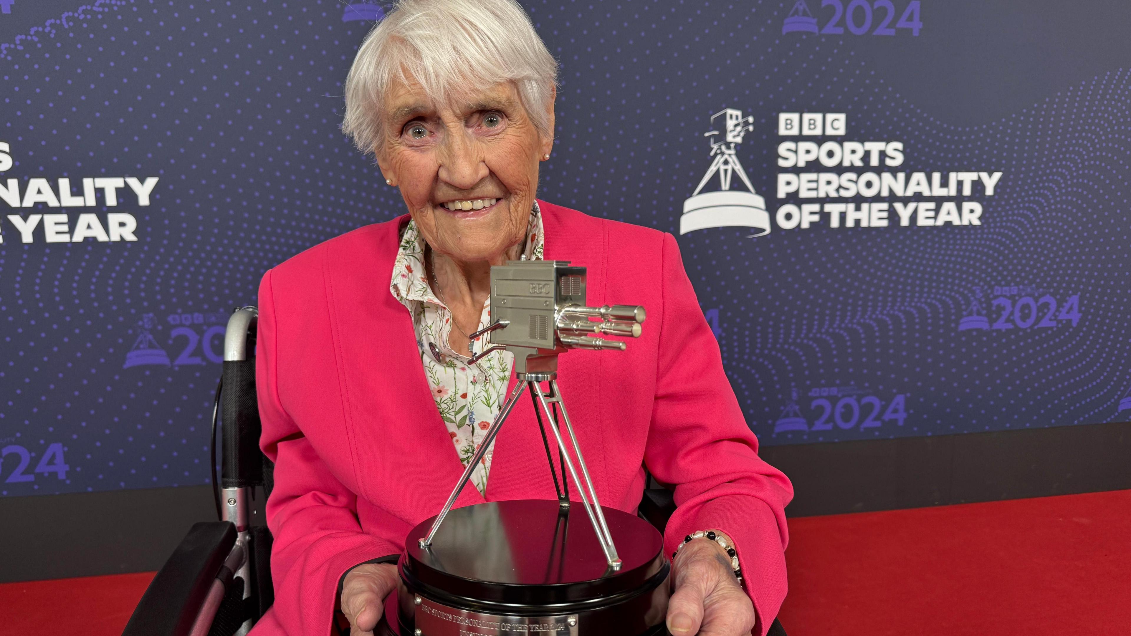 Jean Paton sits in her wheelchair looking directy at the camera, she has white, short hair and wears a bright fuschia pink blazer, buttoned up with a white collared shirt which has a green and pink floral print on it. In the background is a purple sign reading 'BBC Sports Personality of the Year' and Ms Paton is on the red carpet holding her award