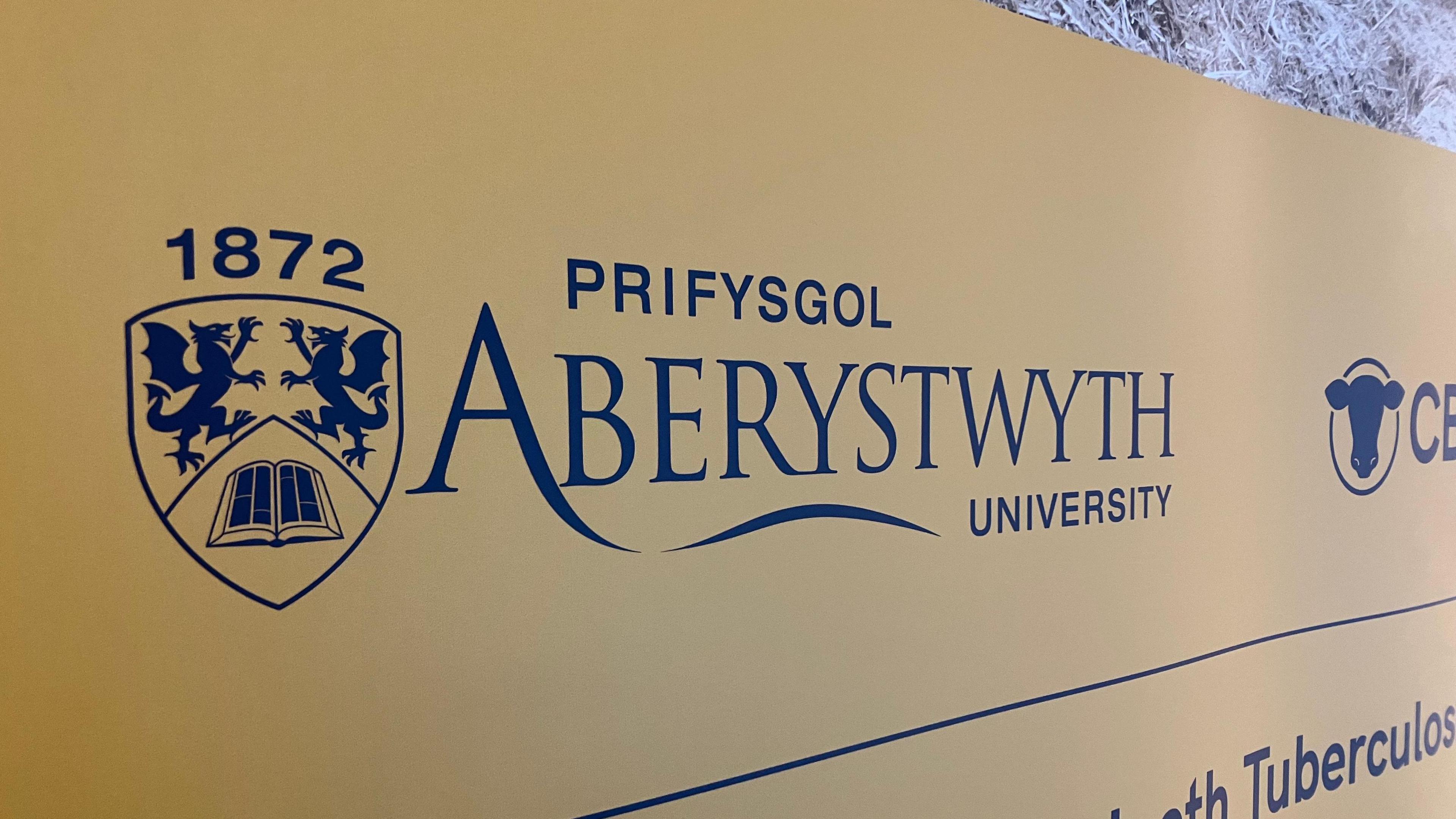 Arwydd Prifysgol Aberystwyth gyda chefndir melyn ac ysgrifen glas.