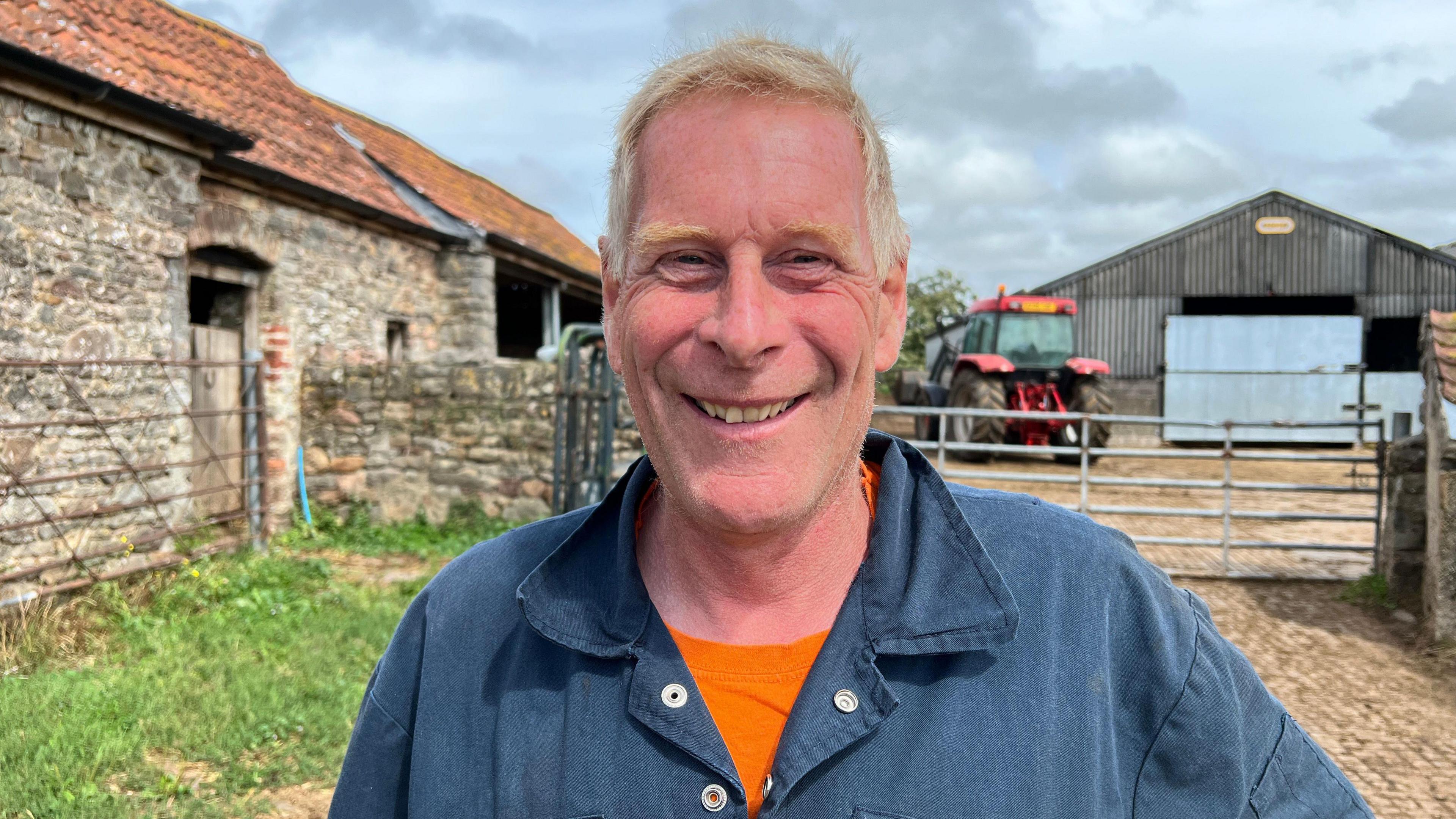 Richard Cornock smiles as he stands on his farm. He is an older man, wearing a blue long-sleeved polo shirt, with a bright orange t-shirt poking out beneath it. He has grey blond hair. A large Cotswold stone building can be seen to his left, and there is a metal gate to his right behind him, which leads to the yard of a corrugated metal barn and a red tractor.