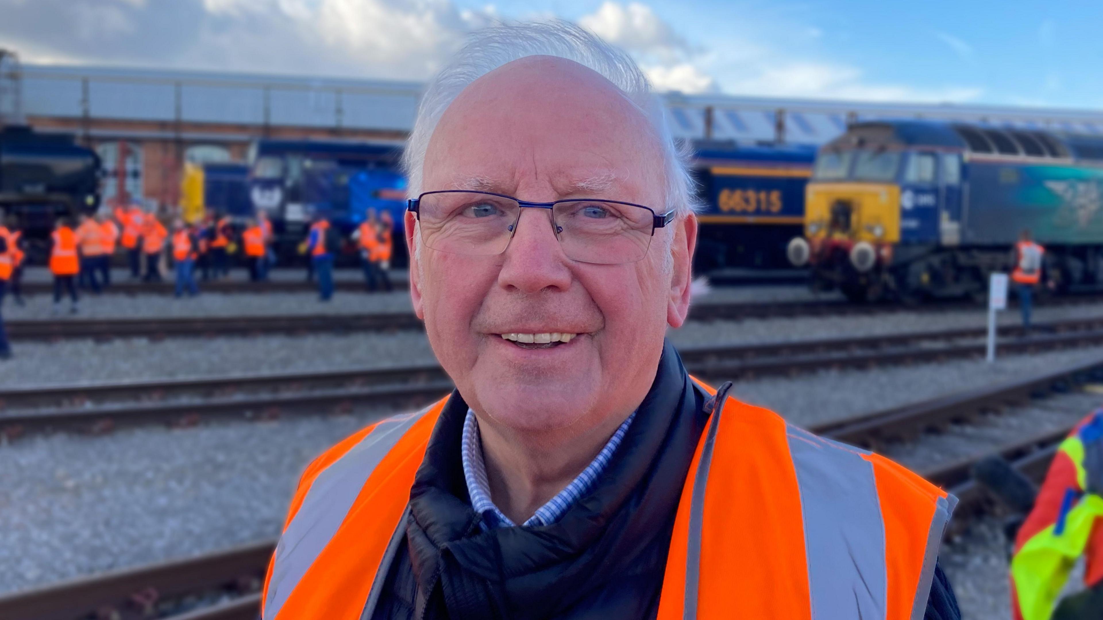 Picture of Pete Waterman on the Alstom rail yard. He is wearing a hi-vis jacket and is stood in front of some historic trains.