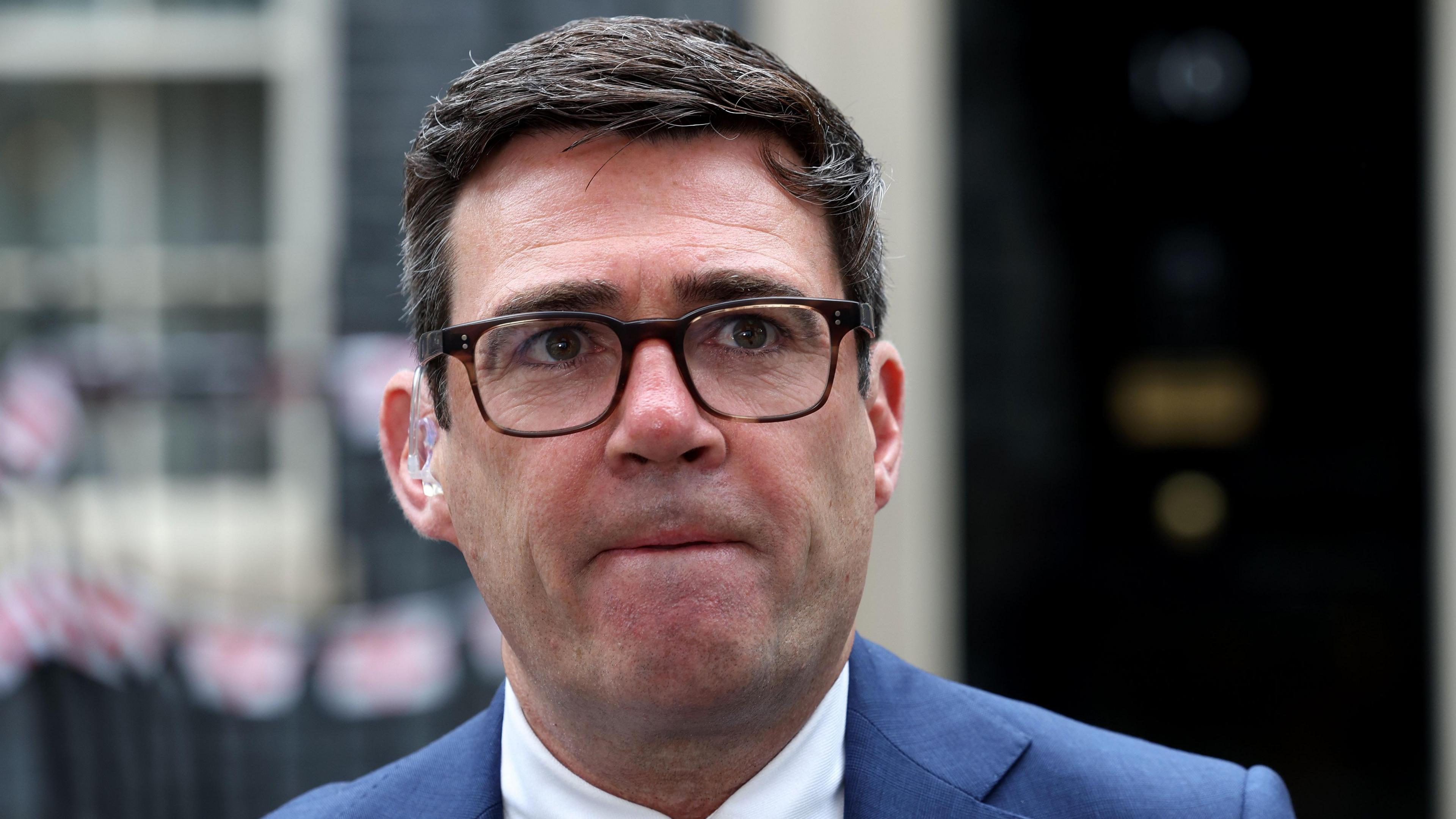 Manchester Mayor Andy Burnham stands in front of No.10, which is blurred out behind him. He wears a blue suit and white shirt with tortoiseshell glasses. He has a clear earpiece fitted to his right ear.