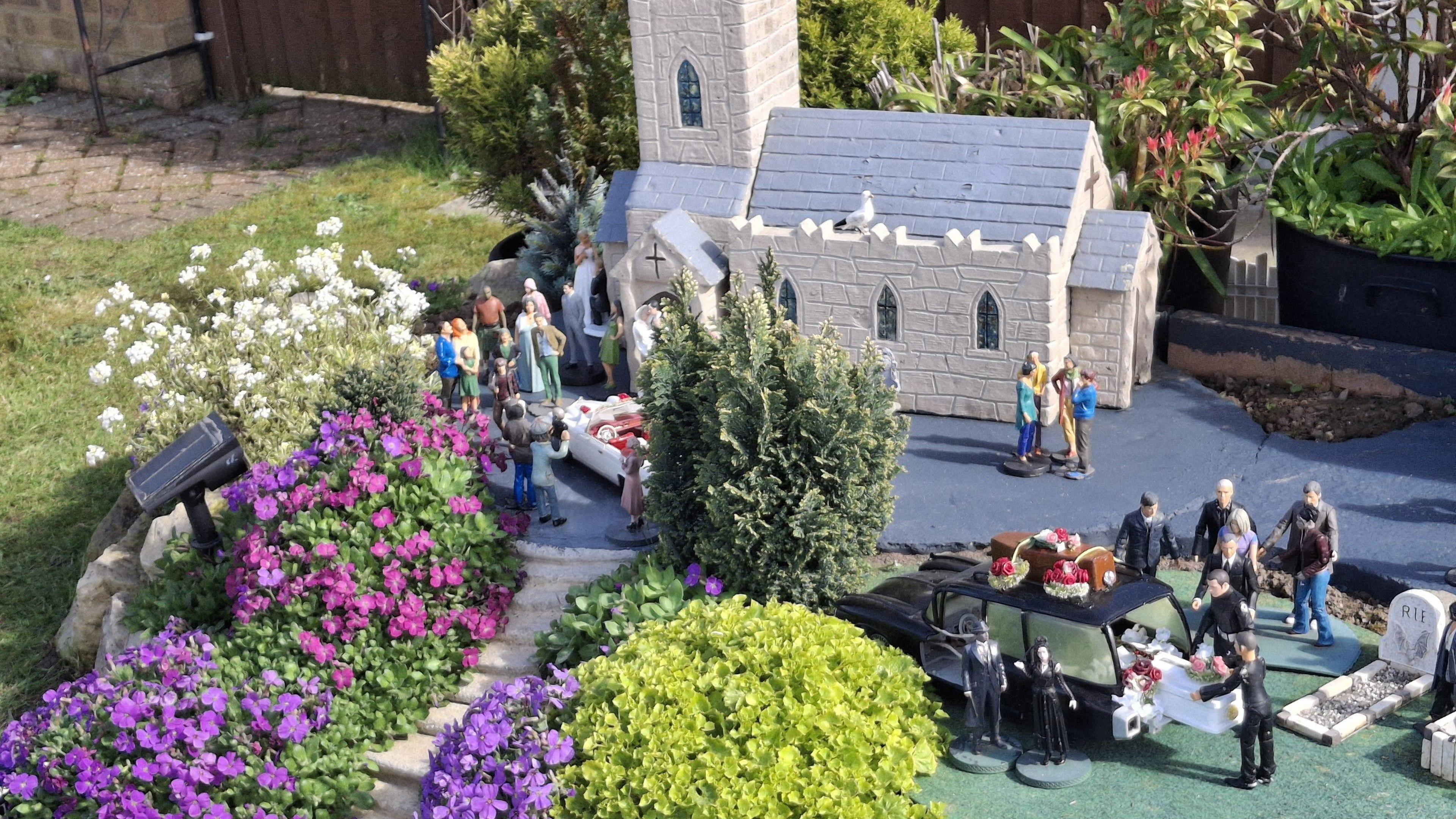 A miniature church as part of a model village. There is a funeral ceremony playing out with the figures including a coffin being lifted from the back of a hearse.