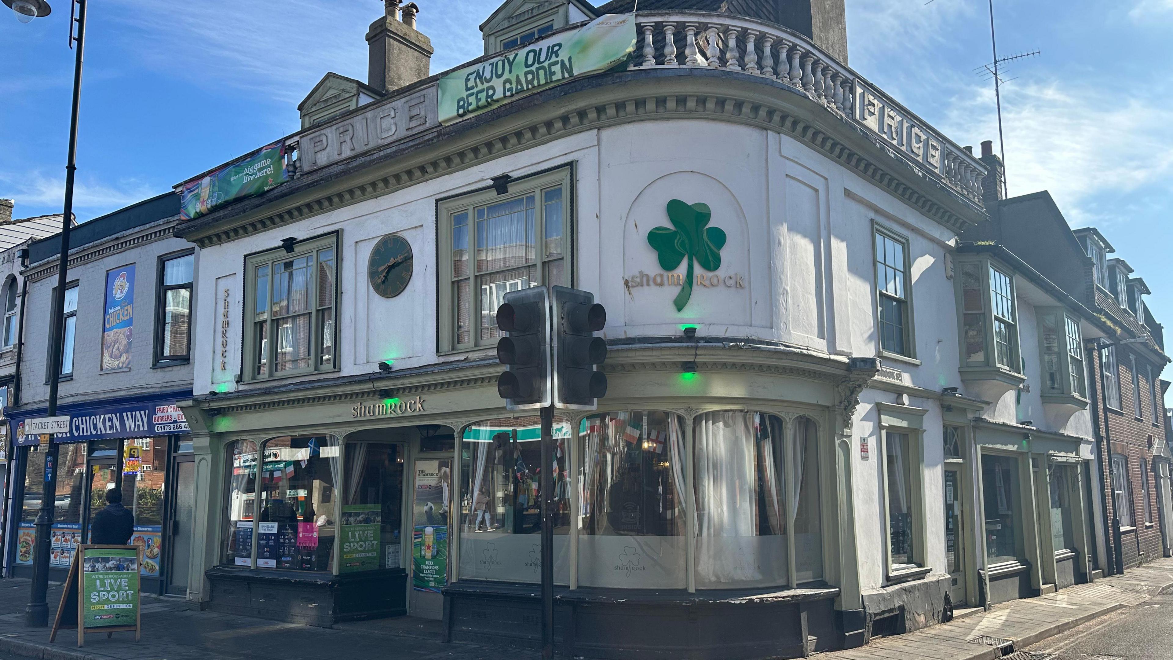 A view of the outside of the Shamrock. It sits on the corner of two streets. On the building there is a green clover leaf. 