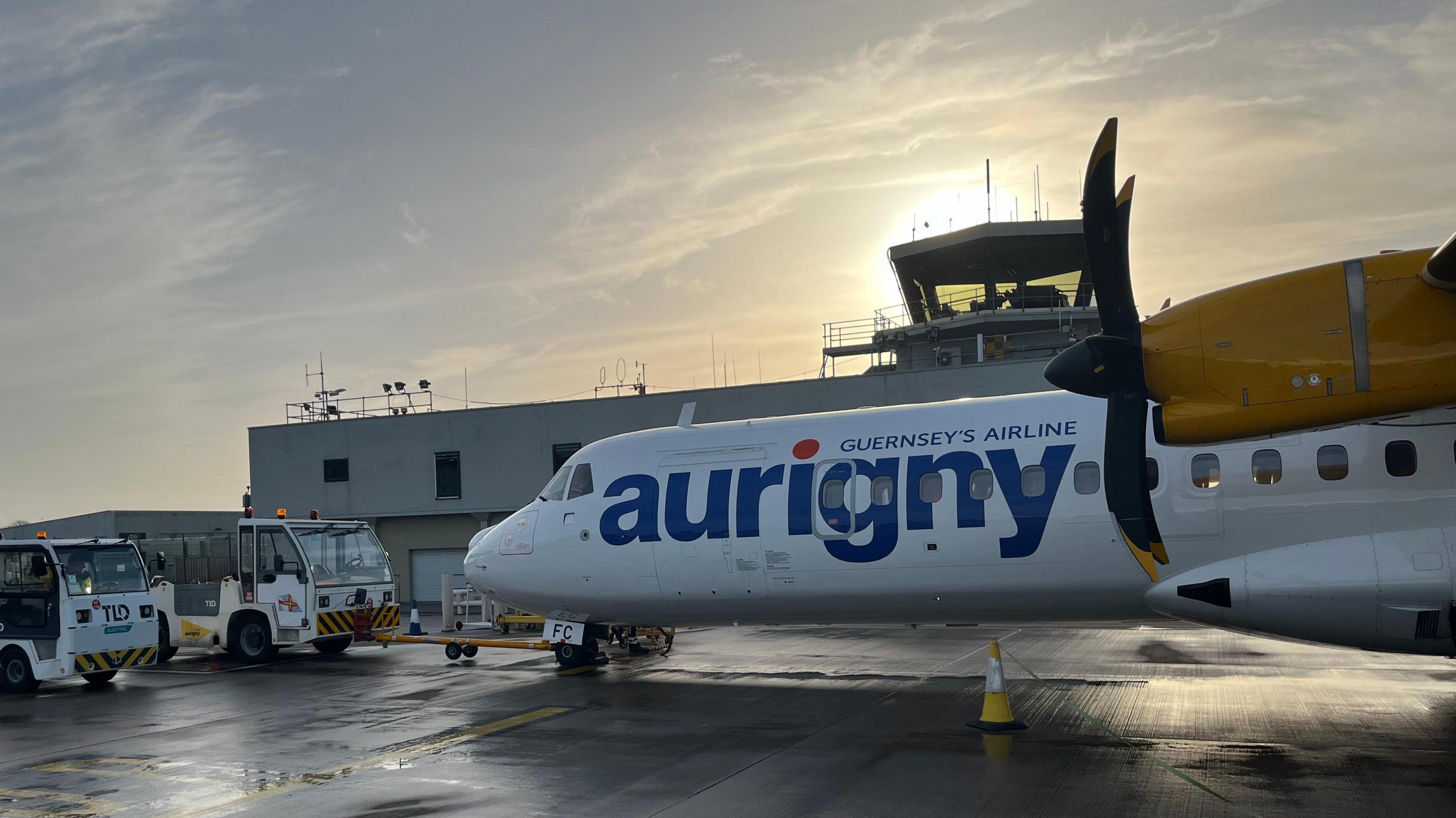 An Aurigny ATR aircraft on the tarmac, with the control tower in the background. The plane is white and yellow with propellers.